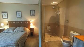 A bedroom with a bed, bedside lamps, and wall art. Adjacent is a bathroom with a shower, toilet, and a small basket.