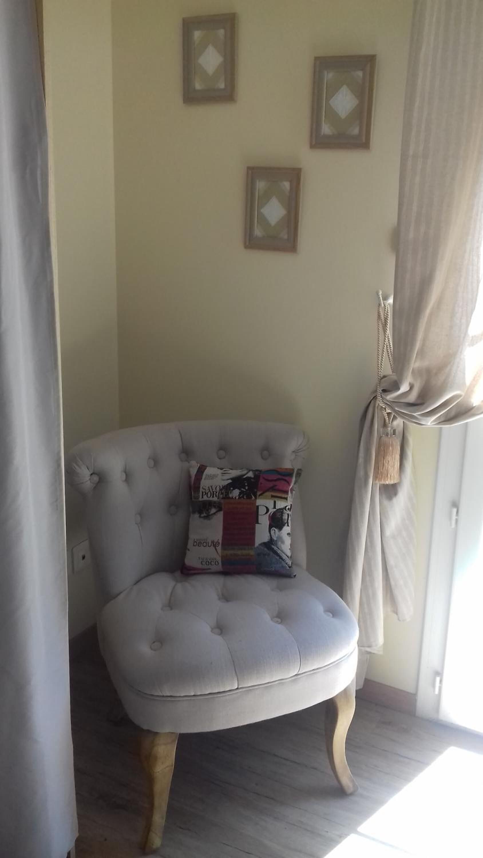 Una habitación de esquina con un sillón capitoné, un cojín estampado y tres cuadros en la pared.