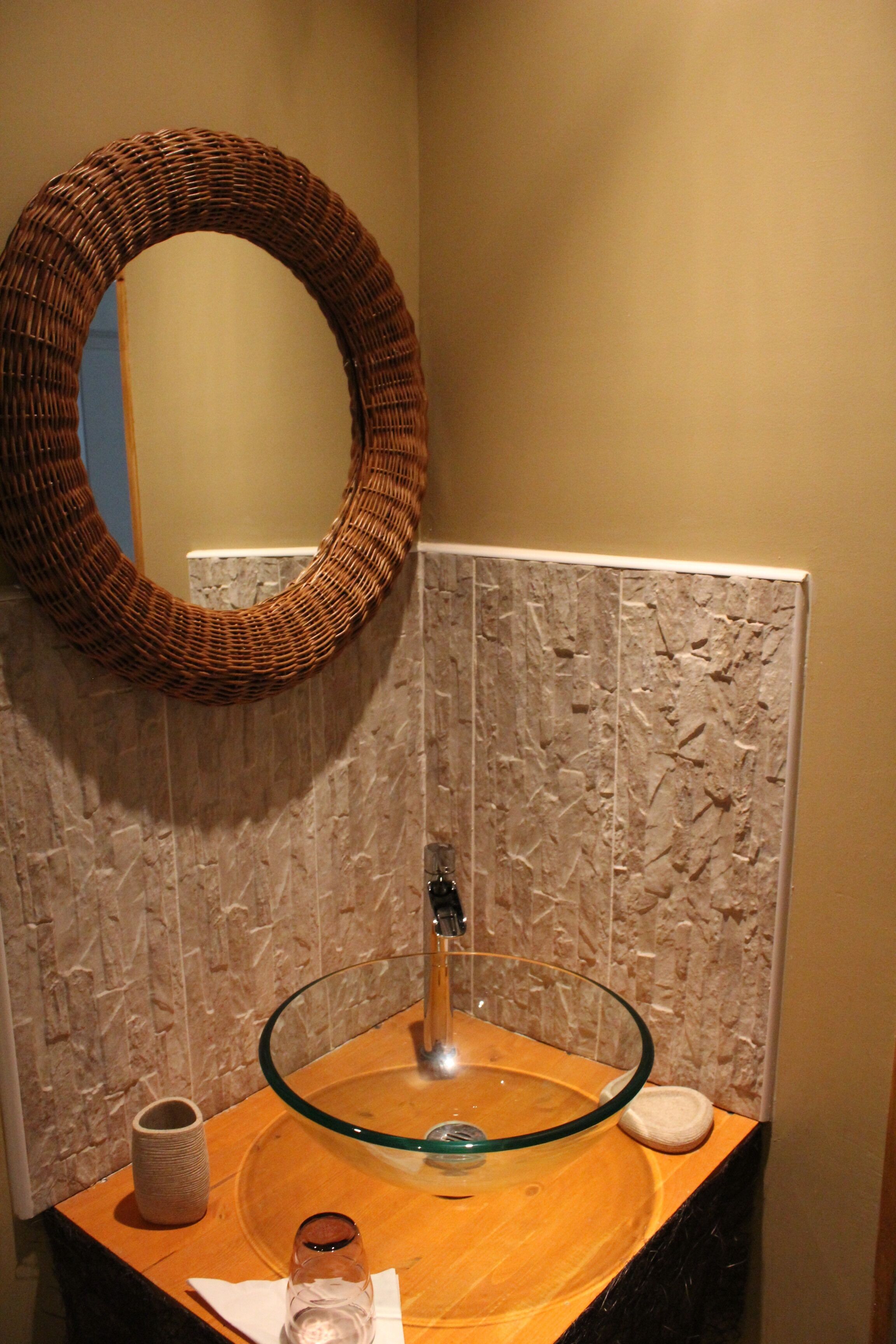 A bathroom with a round glass sink, a wicker mirror, and a marble wall.