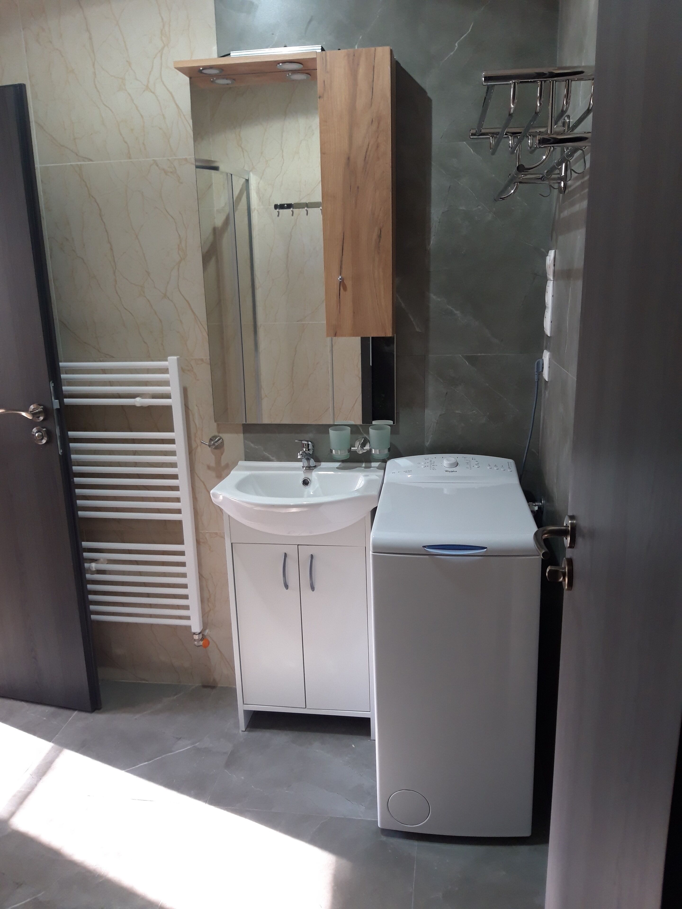 Un baño con lavamanos blanco, un espejo, un mueble de madera, un perchero y una lavadora.