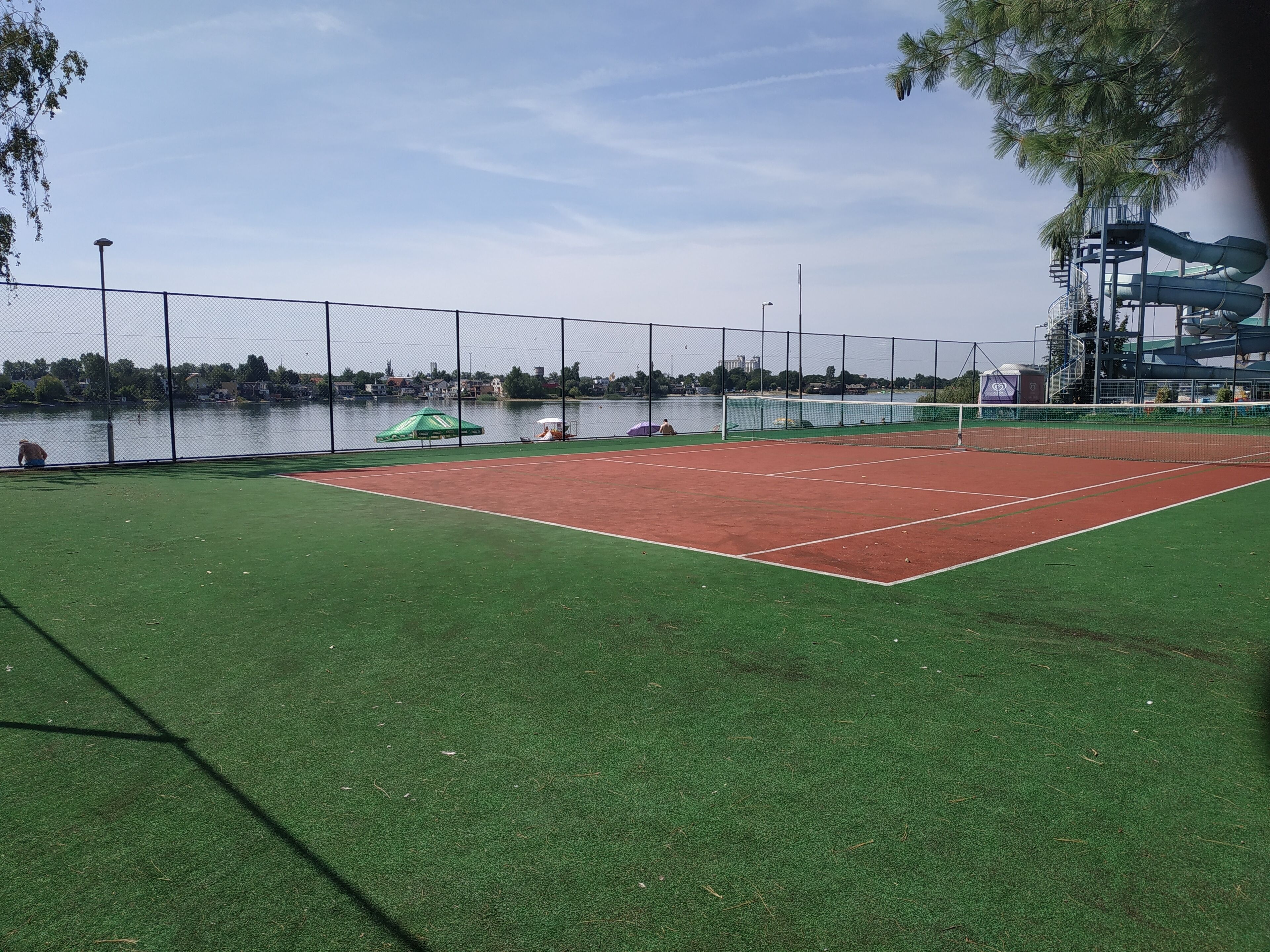 A tennis court with a net, surrounded by a fence, and a body of water in the background.