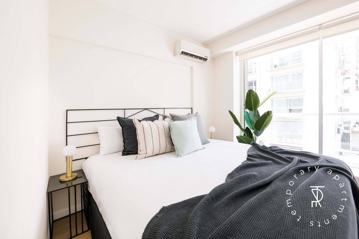 A modern bedroom with a bed, bedside table, lamp, and a window with a view of buildings.