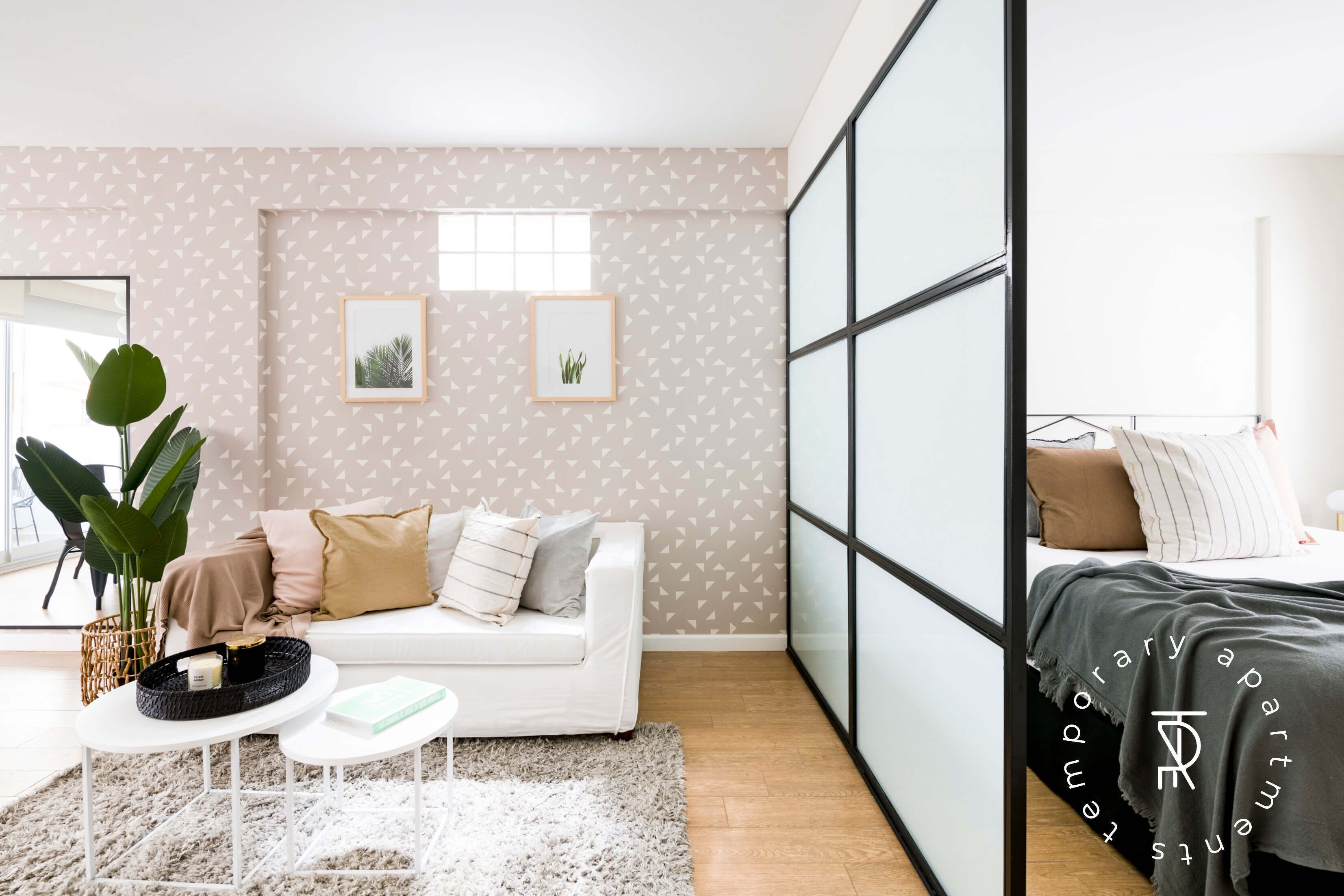 A modern bedroom with a white sofa, a small table, a large plant, and a bed with a grey blanket.