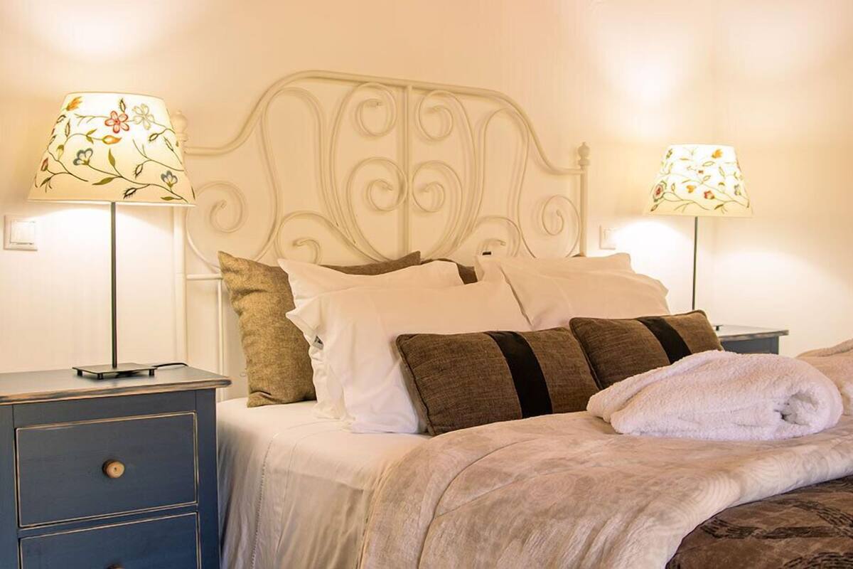 A neatly made bed with white and brown pillows, a folded towel, and two bedside lamps with floral patterns.