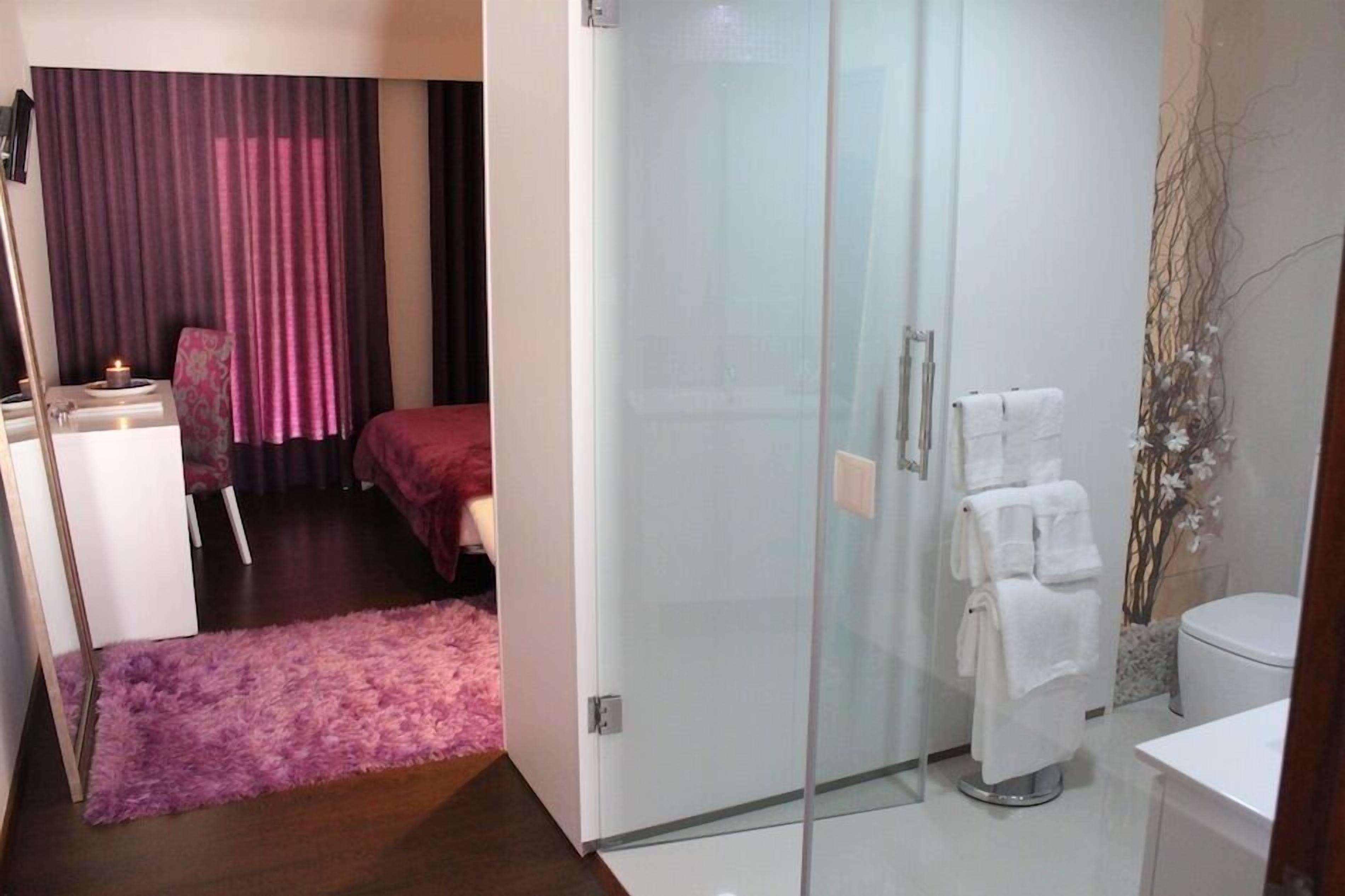 Habitación de hotel con cama, cabina de ducha de vidrio, lavamanos blanco y alfombra morada.