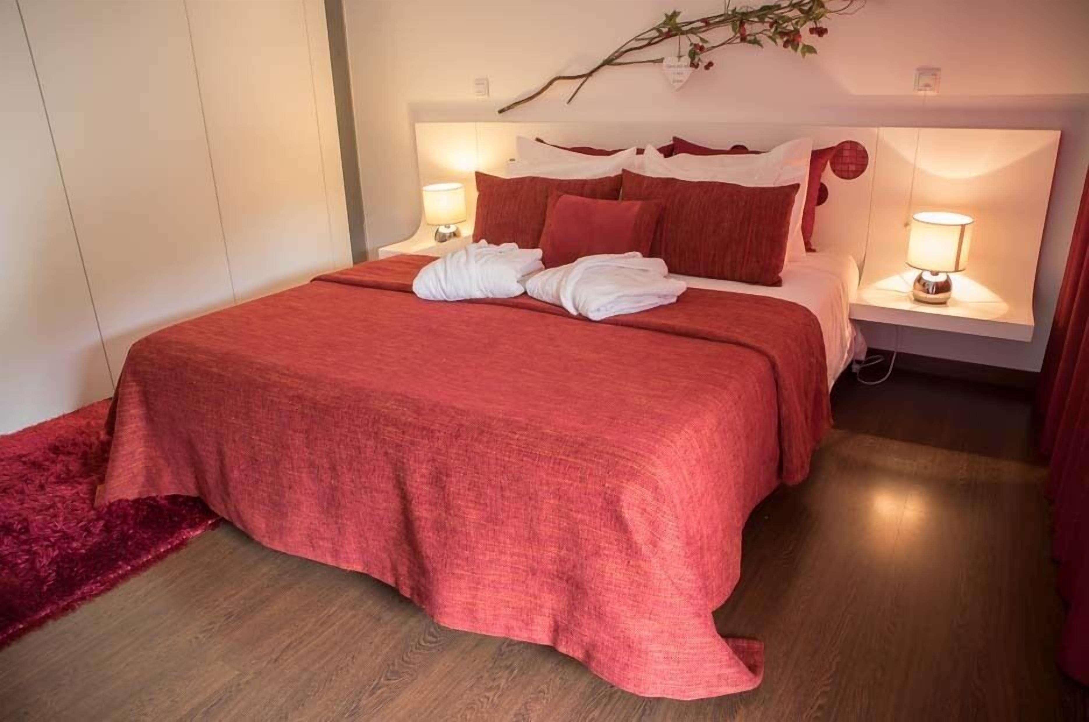 Una cama bien hecha con una colcha roja y almohadas blancas, dos lámparas de noche y una rama decorativa en la cabecera.