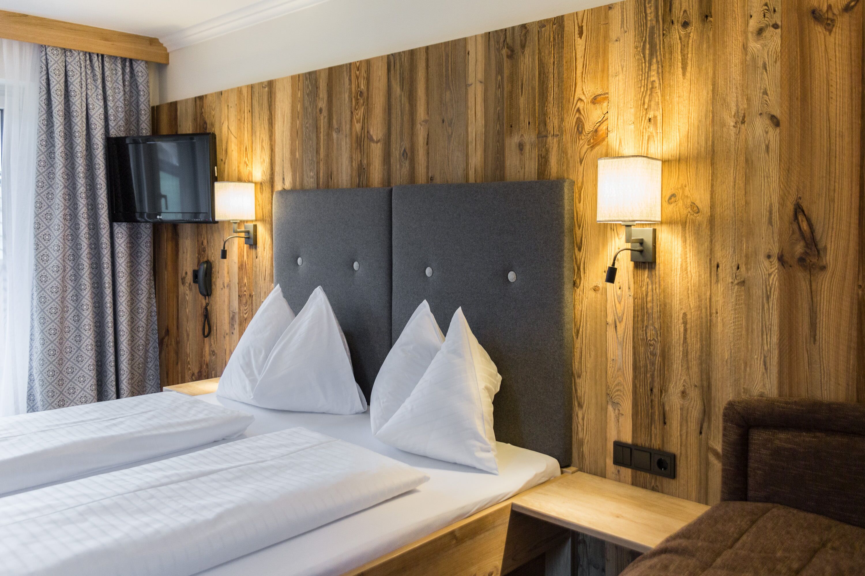 Habitación de hotel con pared de madera, cama con ropa blanca, dos lámparas de noche y televisor montado en la pared.