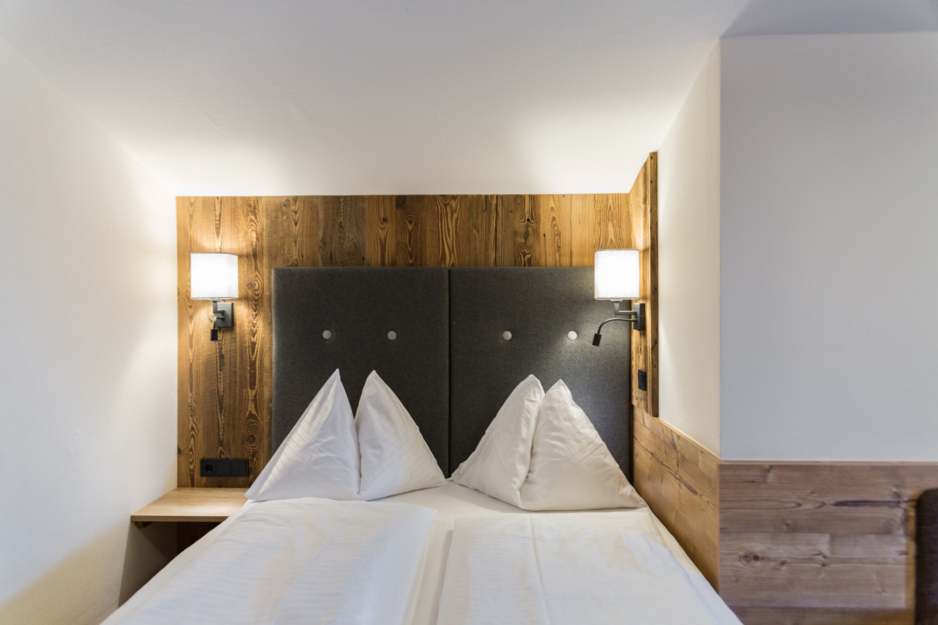 Una habitación de hotel con cabecera de madera, dos lámparas de noche y una cama bien hecha con sábanas blancas.
