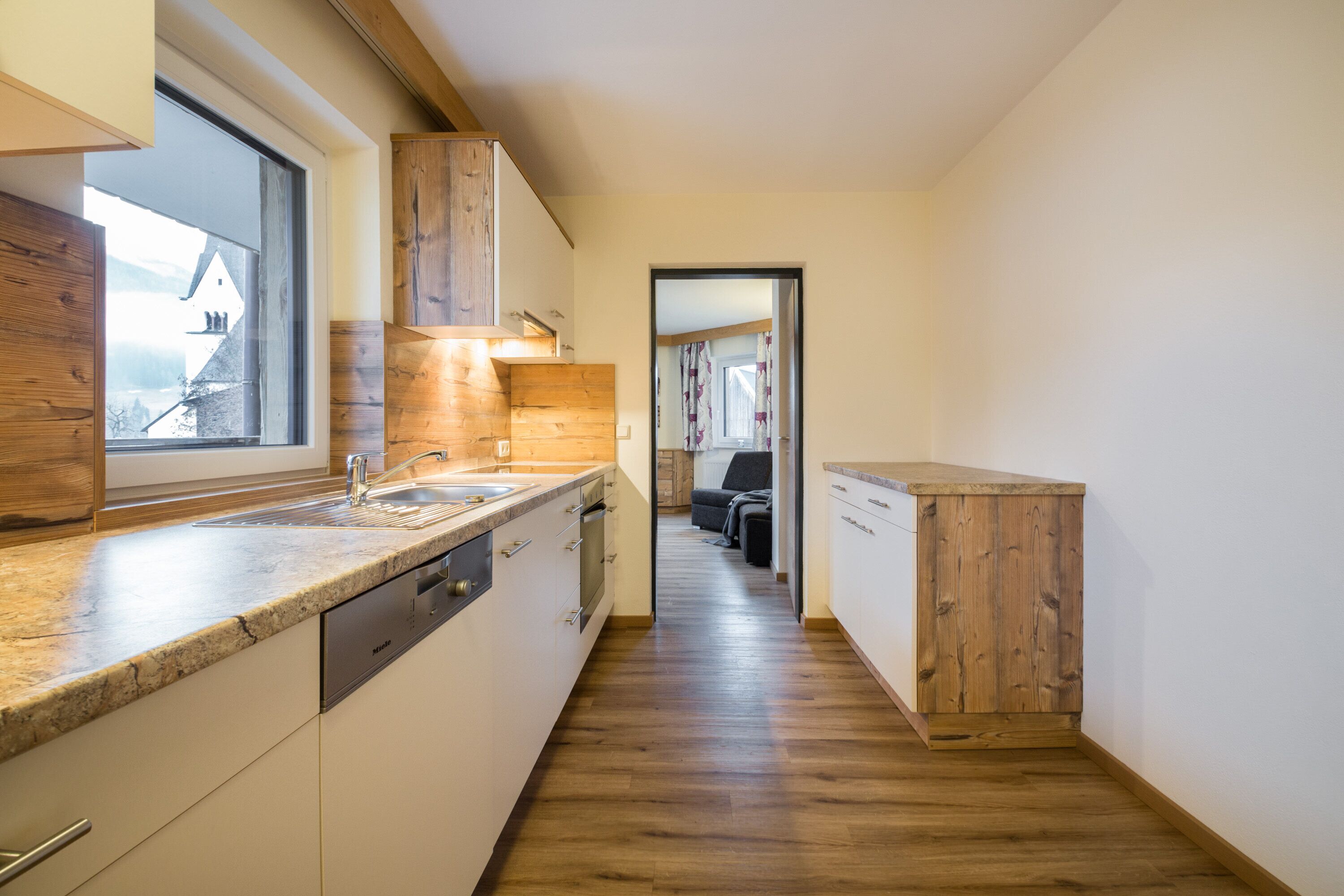 A narrow kitchen with wooden cabinets, a built-in dishwasher, and a window overlooking a house.