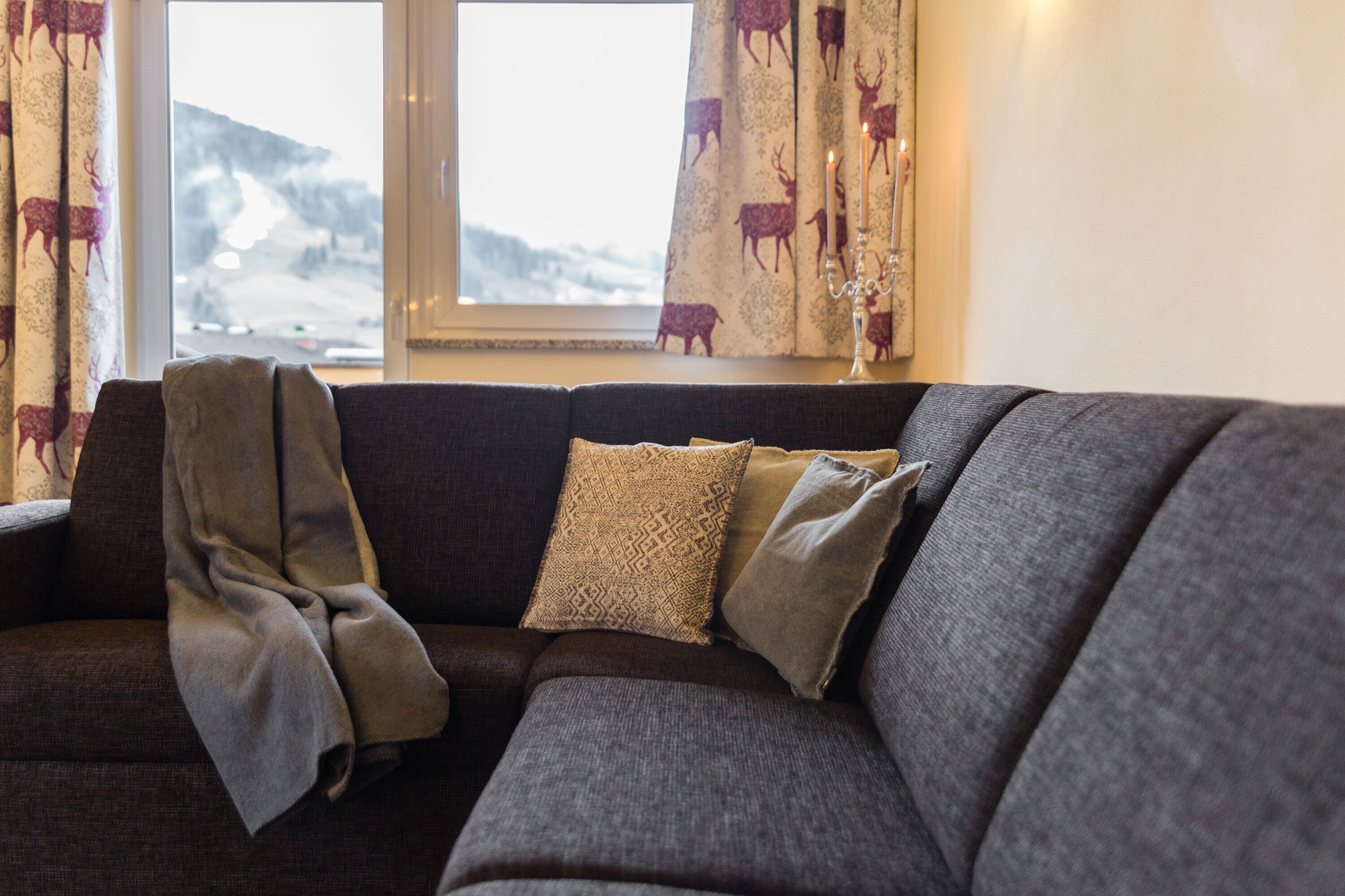 A dark grey sofa with patterned cushions, a blanket draped over it, and a window with curtains featuring a deer pattern.