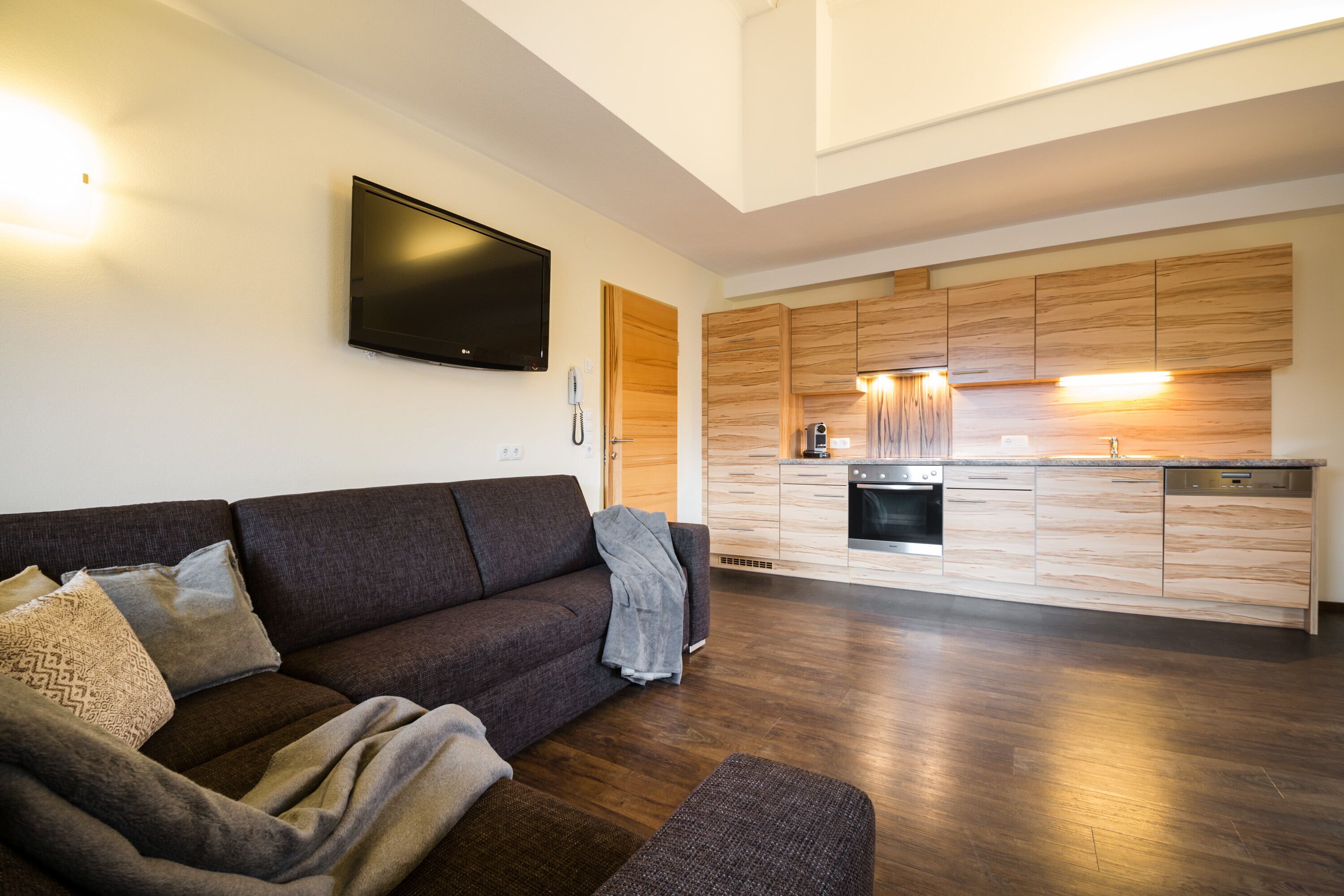 A modern living room with a dark brown sofa, a flat-screen TV mounted on the wall, and a wooden kitchen with built-in appliances.