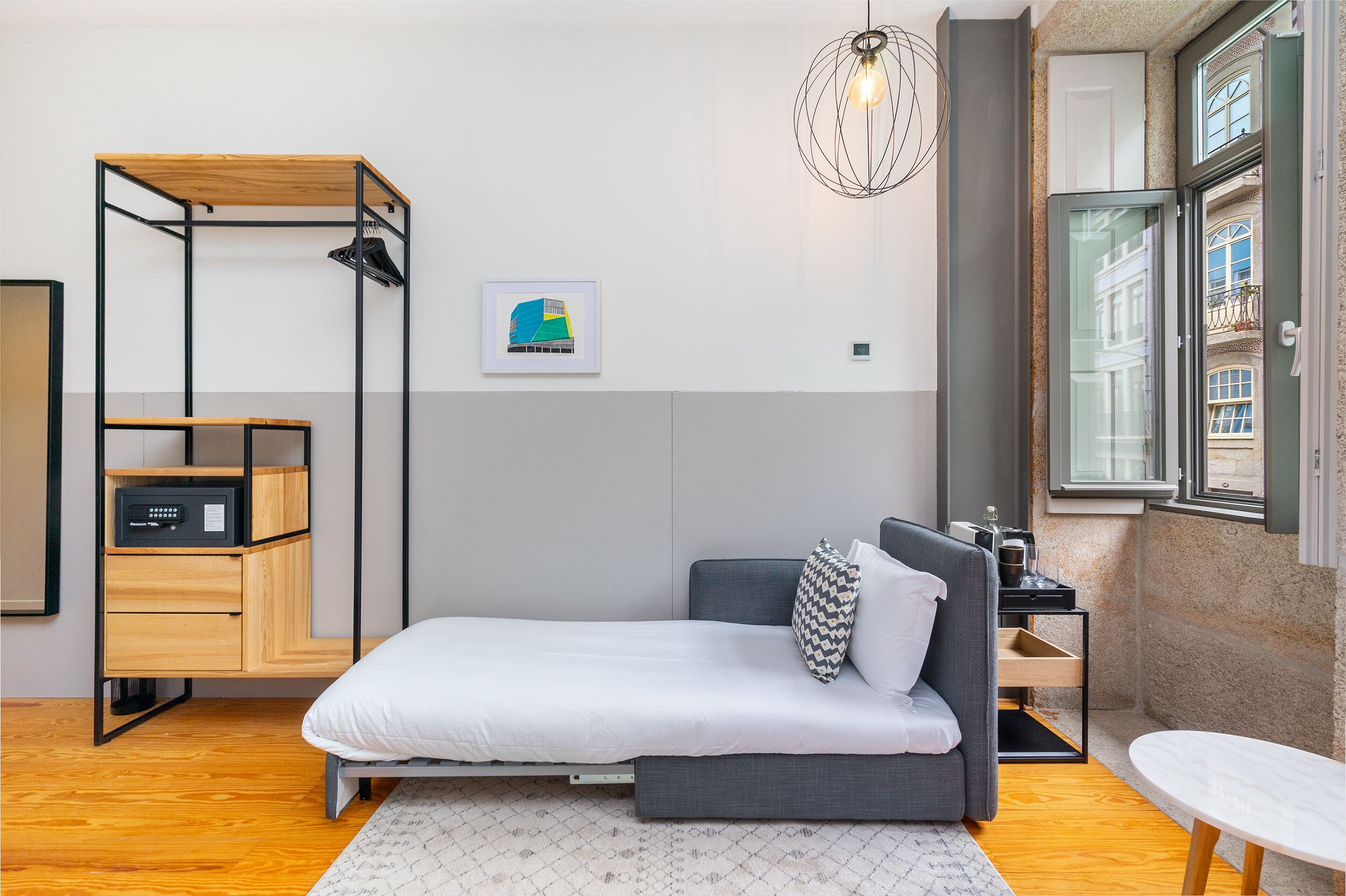 A compact bedroom with a bed, a small table, a hanging light fixture, and a window with a view of buildings.