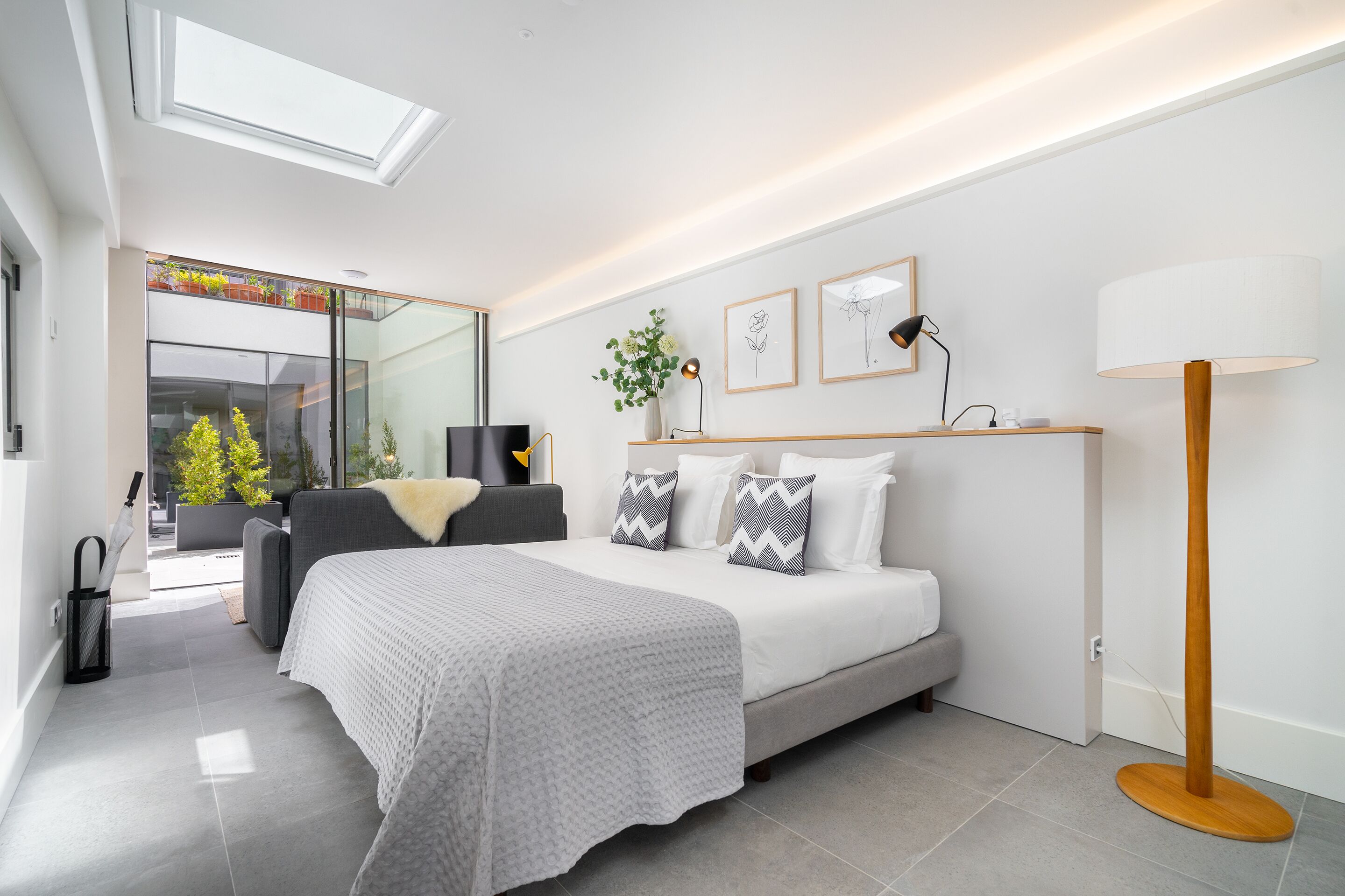 A modern bedroom with a large bed, bedside tables, a floor lamp, and a view of a balcony with plants.