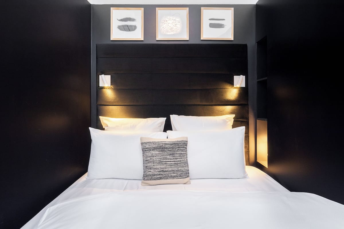 A neatly made bed with white linens and a patterned pillow, a dark headboard with wall-mounted lights, and three framed artworks above the bed.