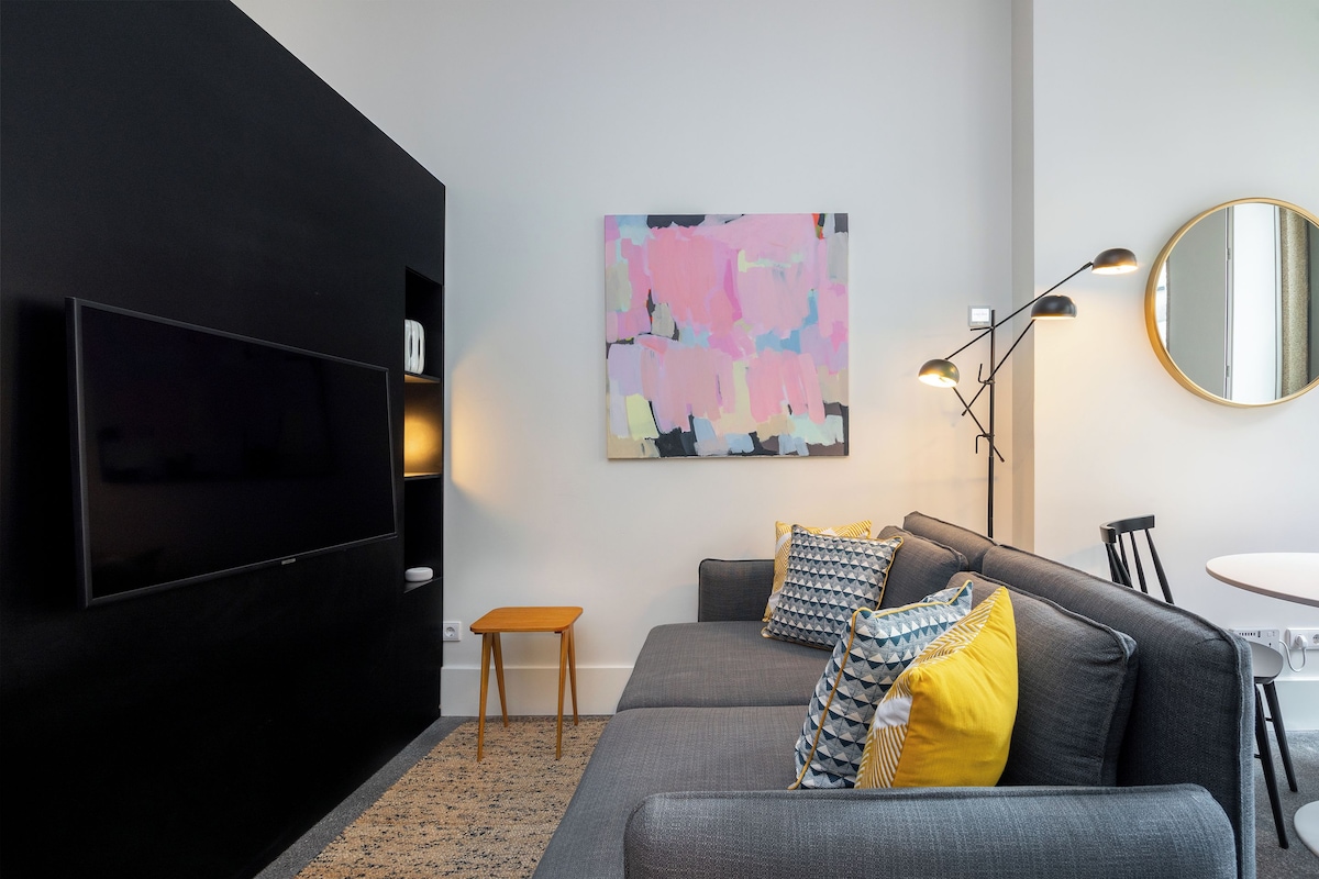 A modern living room with a flat-screen TV, a gray sofa with patterned cushions, a small wooden side table, and a colorful abstract painting on the wall.