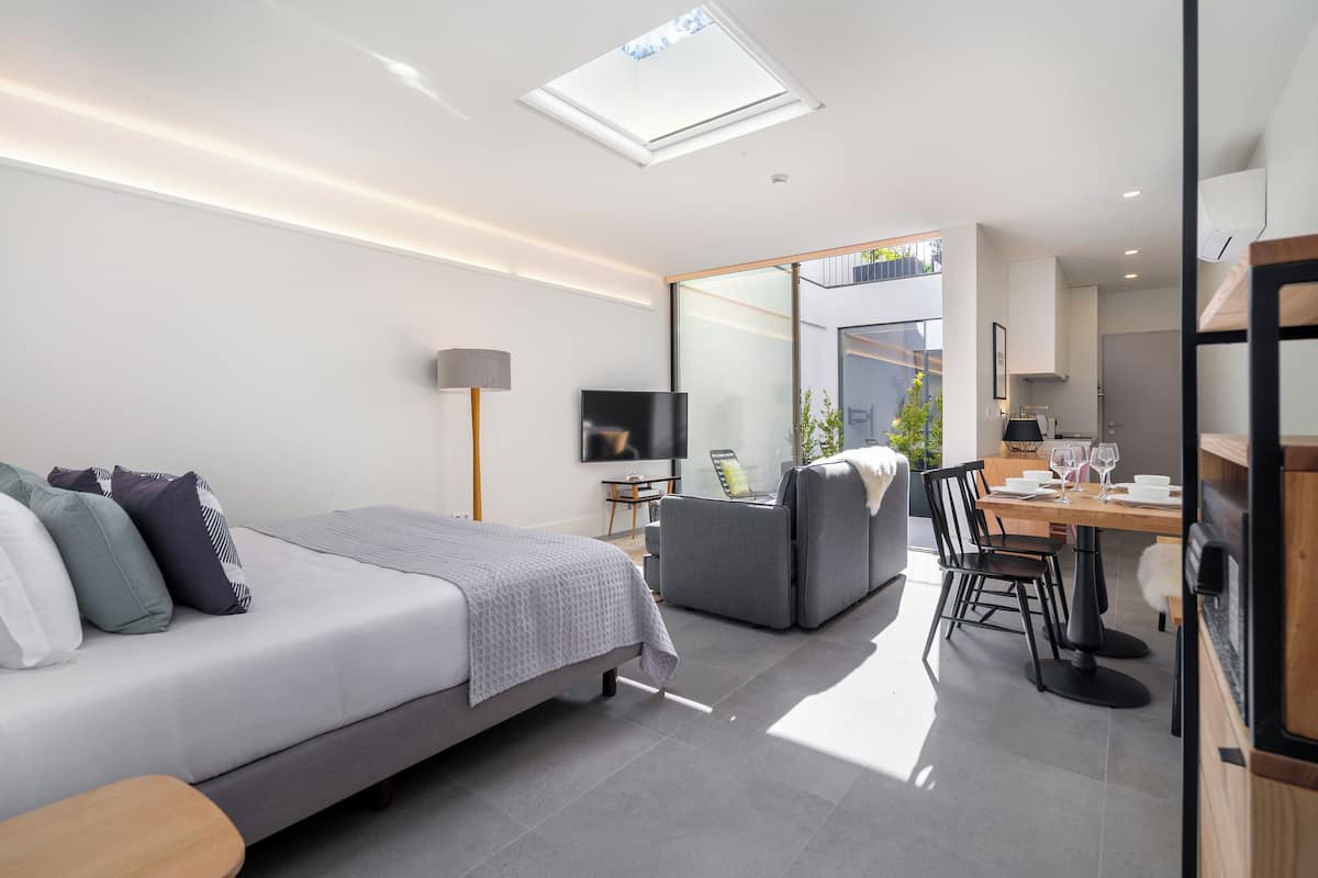 A modern bedroom with a bed, bedside table, and a TV. There is a dining area with a table and chairs.