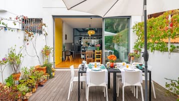 Un patio con mesa lista para comer, rodeado de macetas con plantas y un paraguas grande.