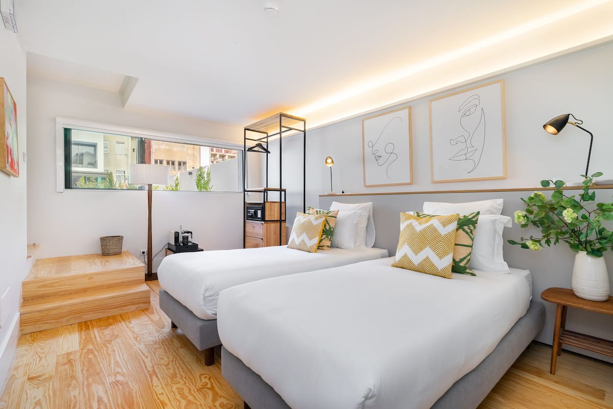 A modern hotel room with two beds, a wooden floor, a window with a view, a desk with a lamp, and artwork on the wall.