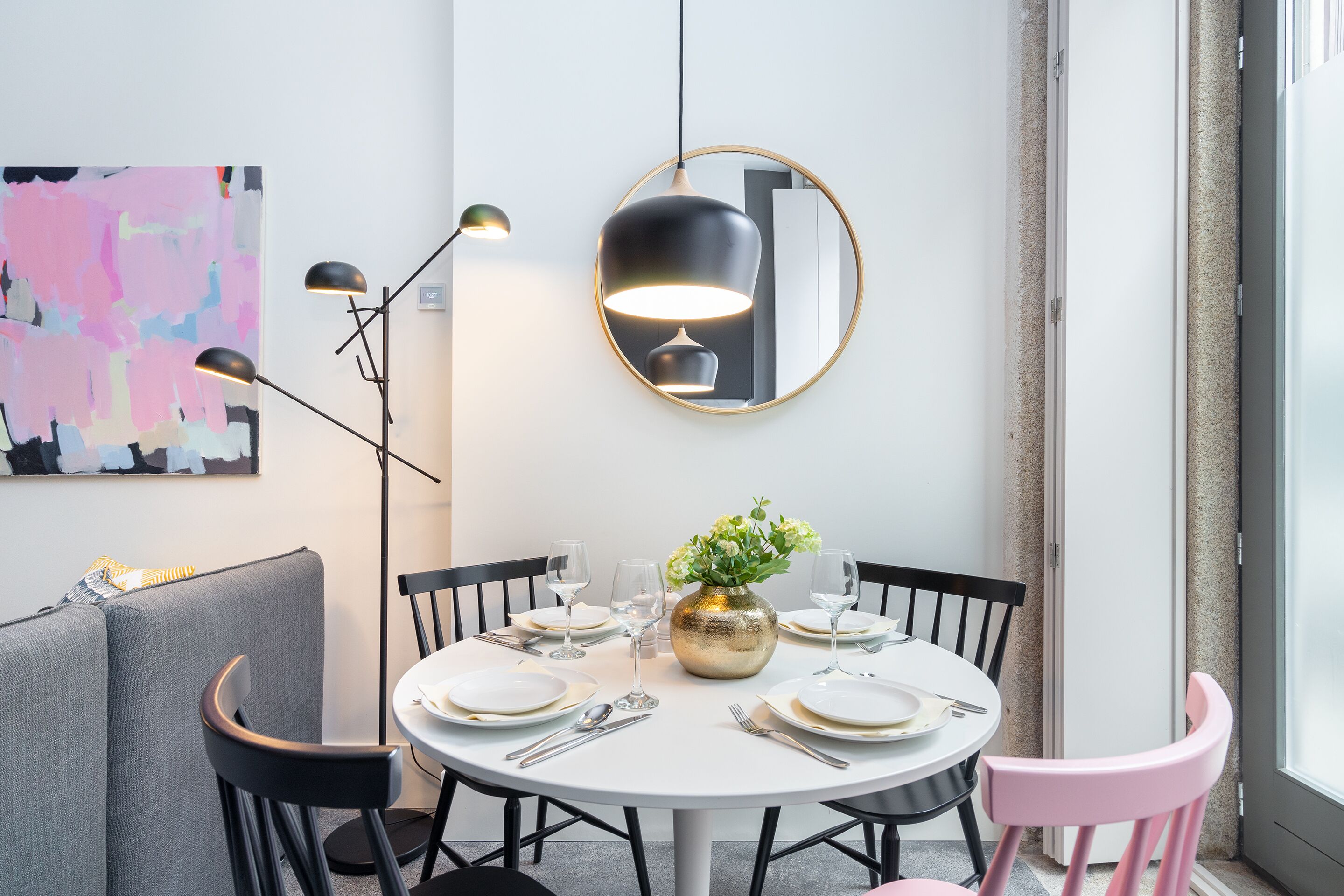 A dining area with a round table set for four, surrounded by black chairs, a gold vase with flowers, and a modern pendant light.