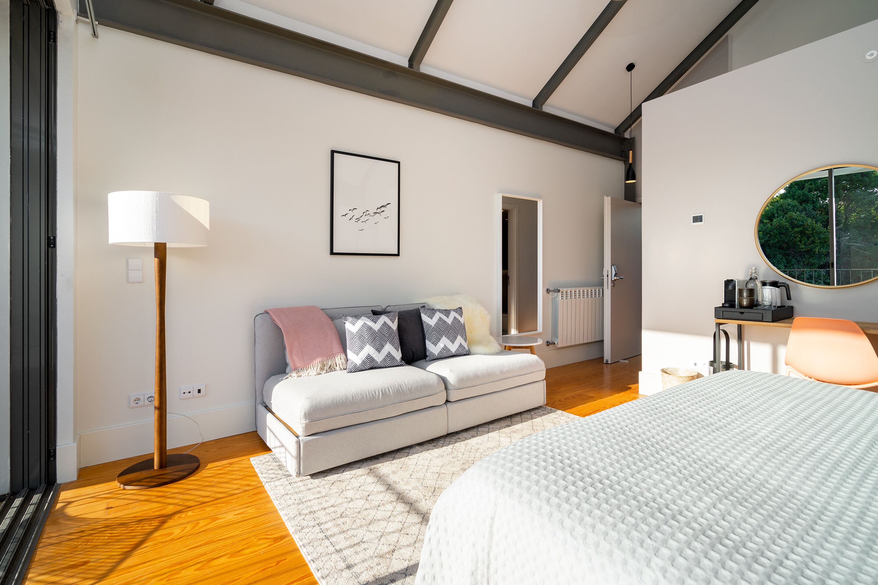 Quarto moderno com cama, sofá, luminária de chão e espelho redondo.