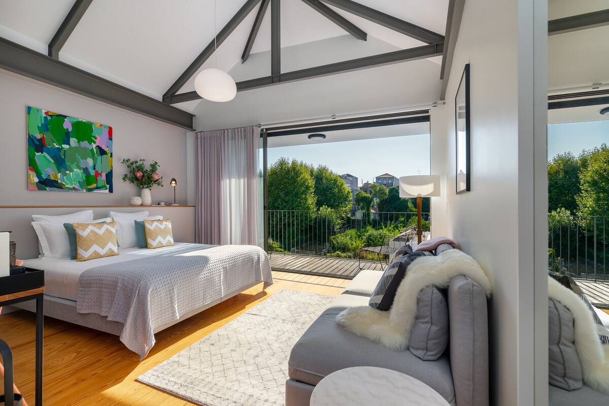 A modern bedroom with a large bed, a sofa, and a balcony with a view of greenery and buildings.