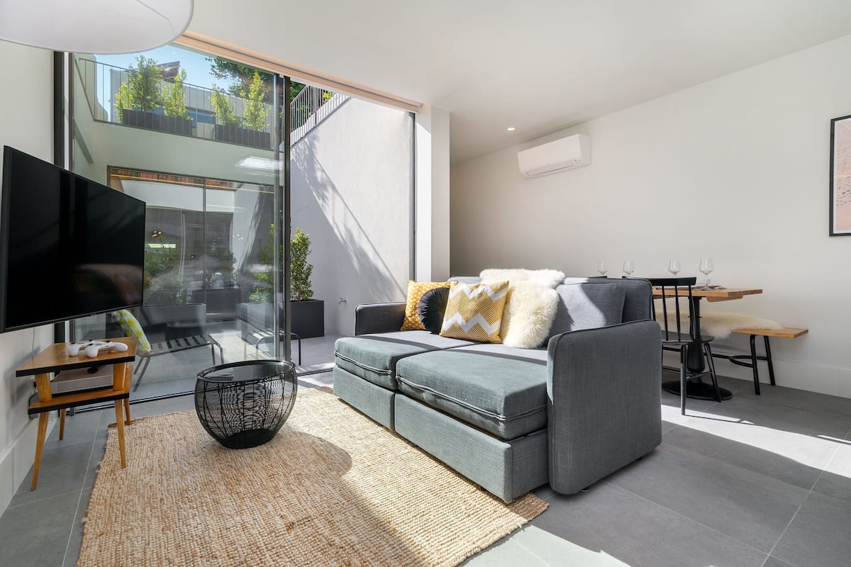 A modern living room with a grey sofa, a flat-screen TV, a dining table, and a large window with a view of greenery.