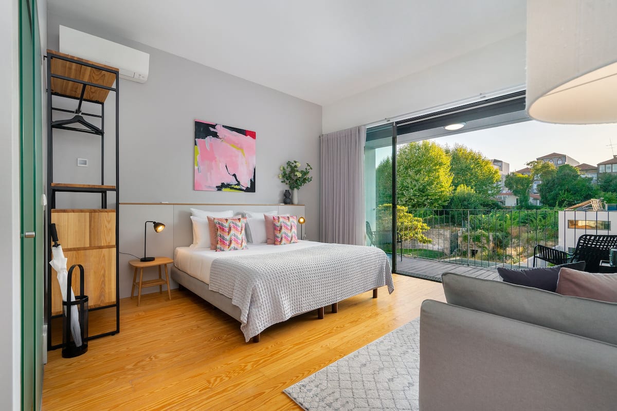 A modern bedroom with a large bed, a wooden wardrobe, a desk, and a balcony with a view of the neighborhood.