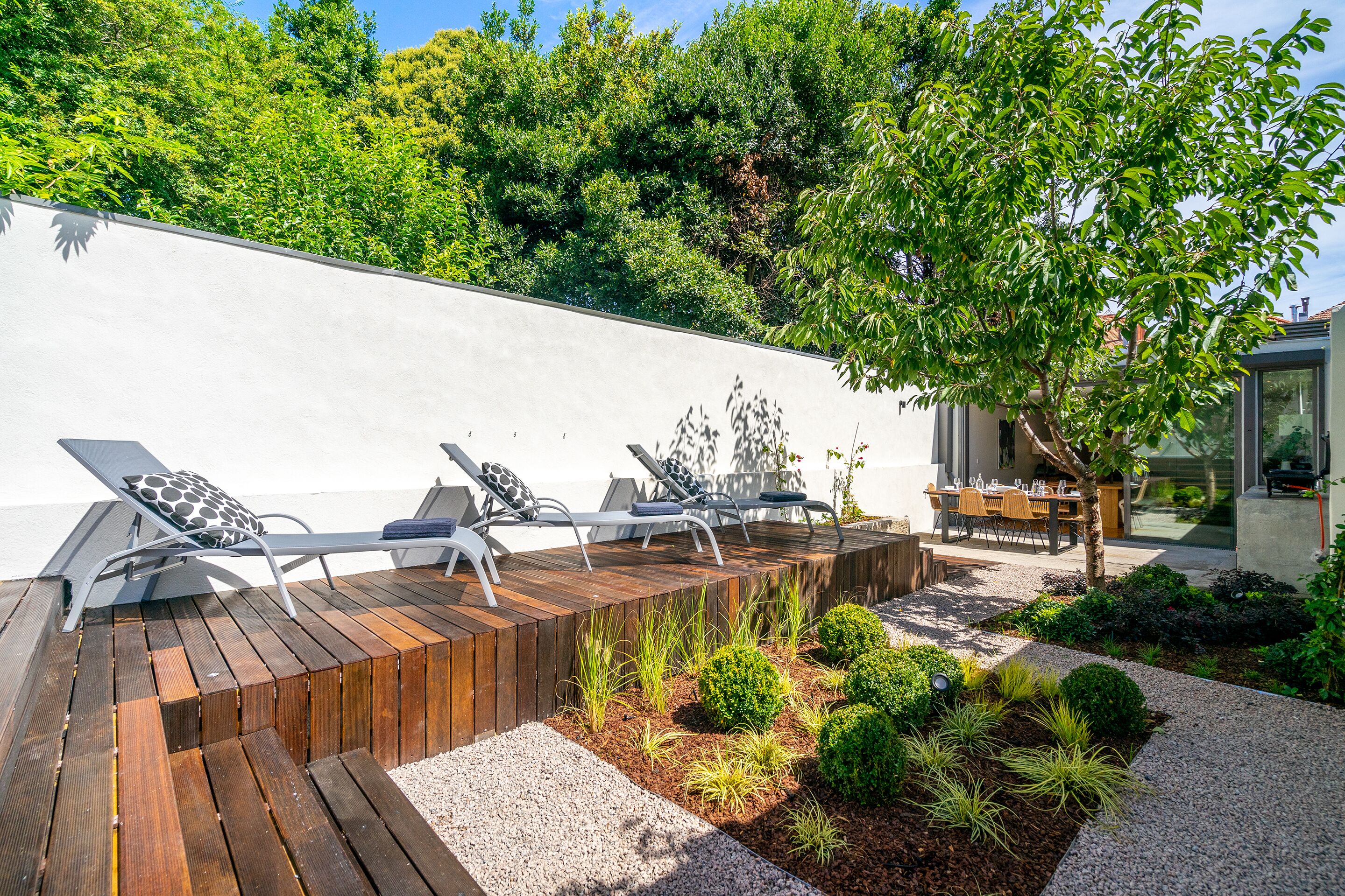 A modern outdoor seating area with wooden deck and lounge chairs, surrounded by landscaped garden beds and a white wall.