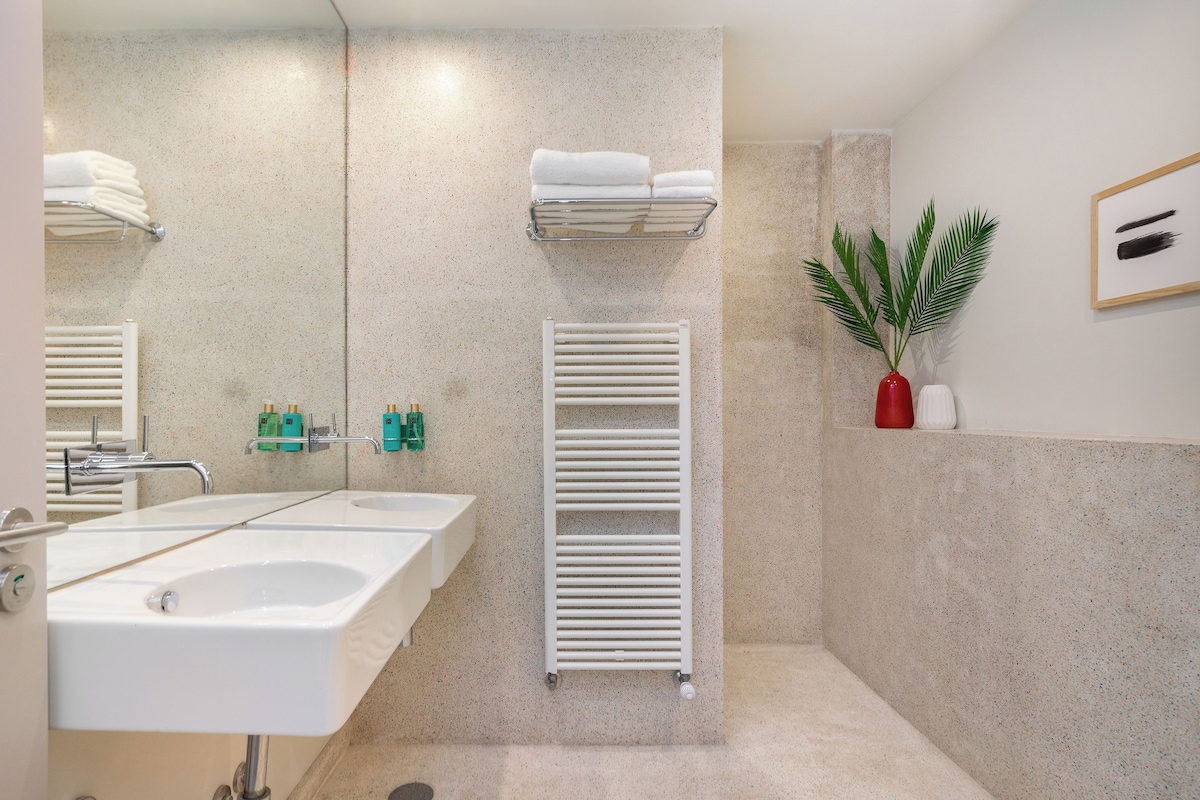 A modern bathroom with two sinks, a towel rack, a radiator, and a potted plant.