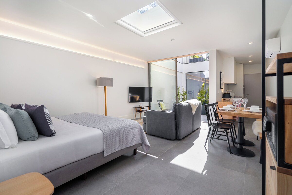 A modern bedroom with a bed, bedside table, and a TV. There is a dining area with a table and chairs.