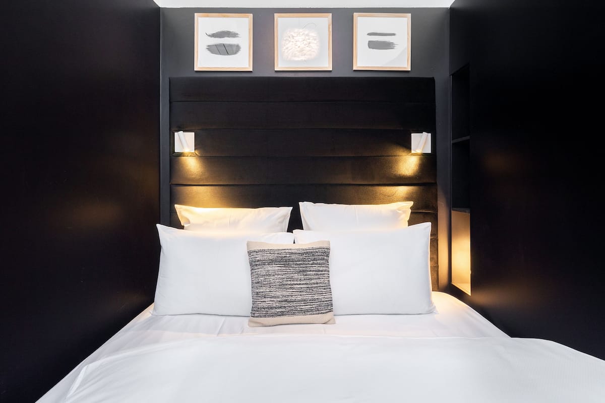 A neatly made bed with white linens and a patterned pillow, a dark headboard with wall-mounted lights, and three framed artworks above the bed.