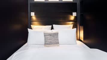 A neatly made bed with white linens and a patterned throw pillow, a dark headboard with wall-mounted lights, and three framed artworks above the bed.