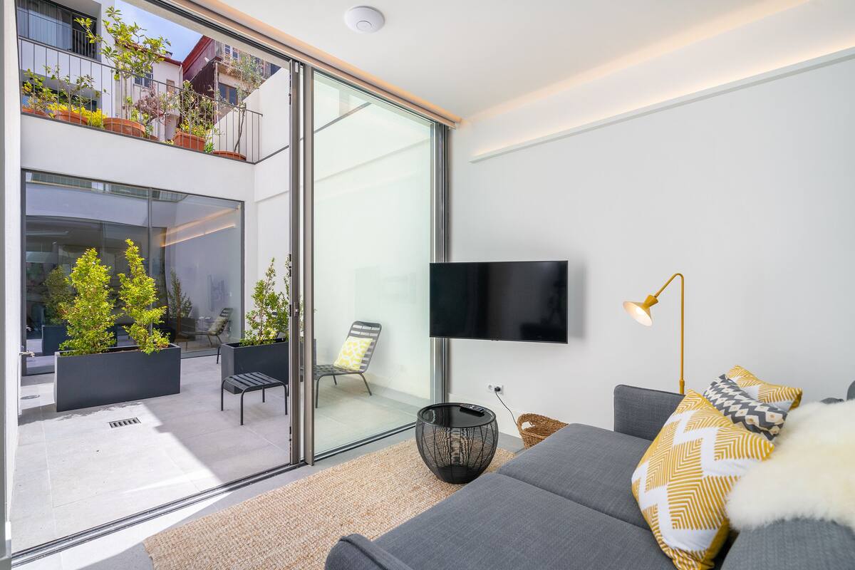 A modern living room with a large flat-screen TV, a sofa with patterned pillows, a floor lamp, and a glass door leading to an outdoor balcony with plants and seating.