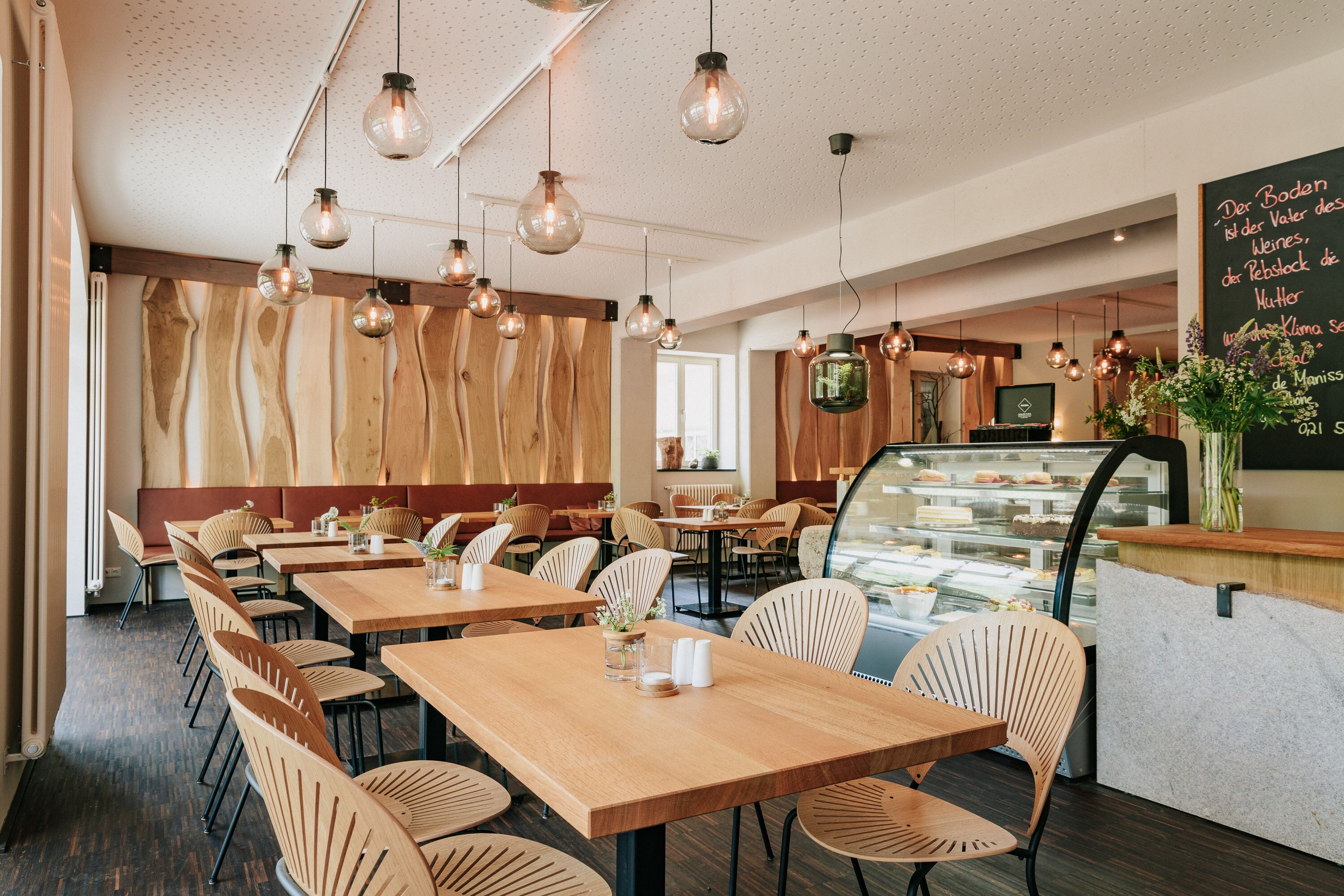 A restaurant interior with wooden tables and chairs, hanging pendant lights, and a chalkboard menu.
