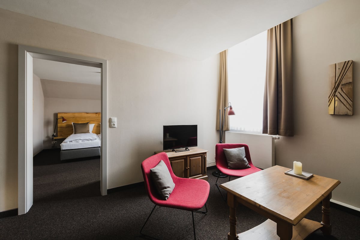 Habitación de hotel con una cama, televisor, mesa de madera, dos sillas rojas y una ventana con cortinas.