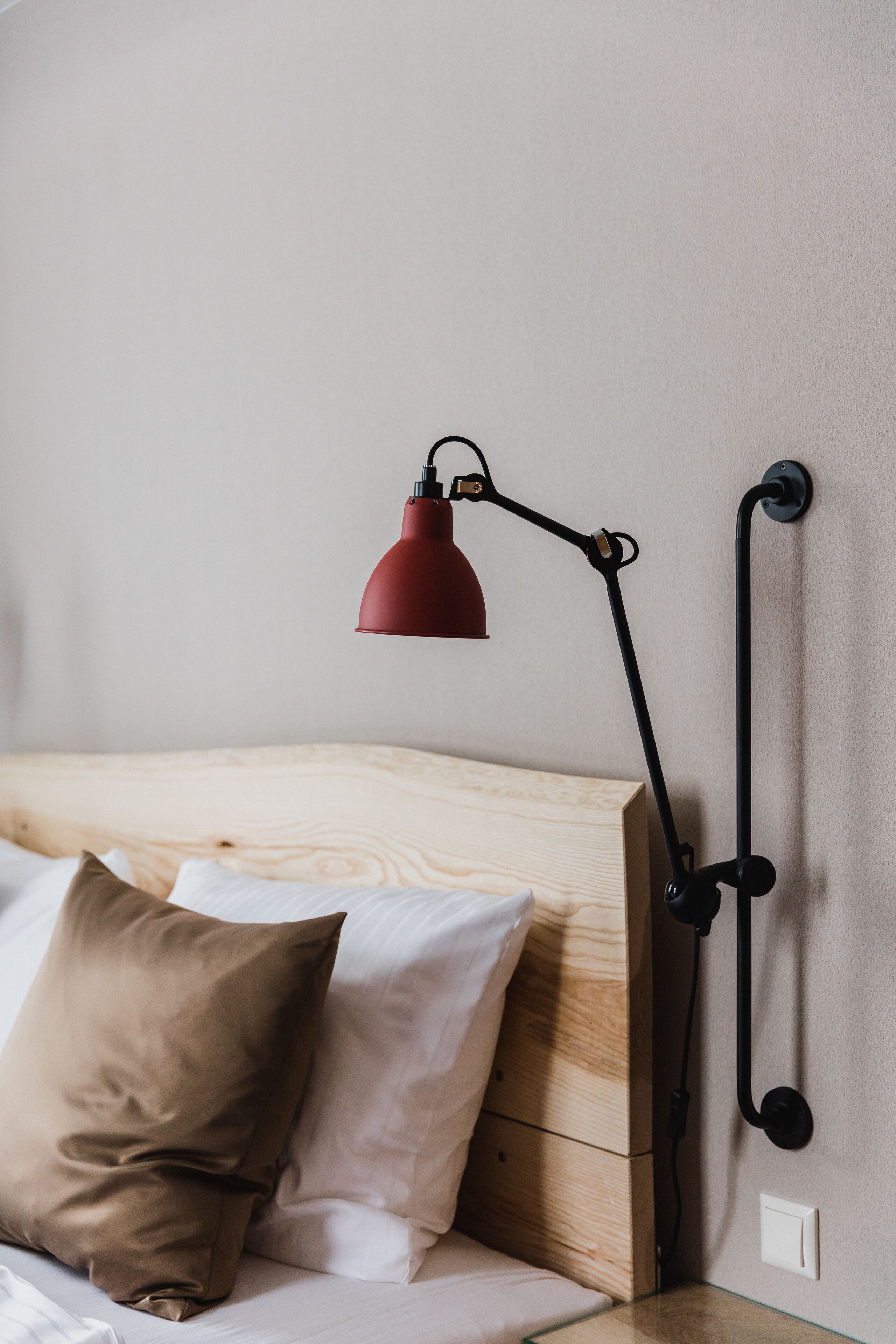 A bed with a wooden headboard, a red desk lamp, and a brown pillow.