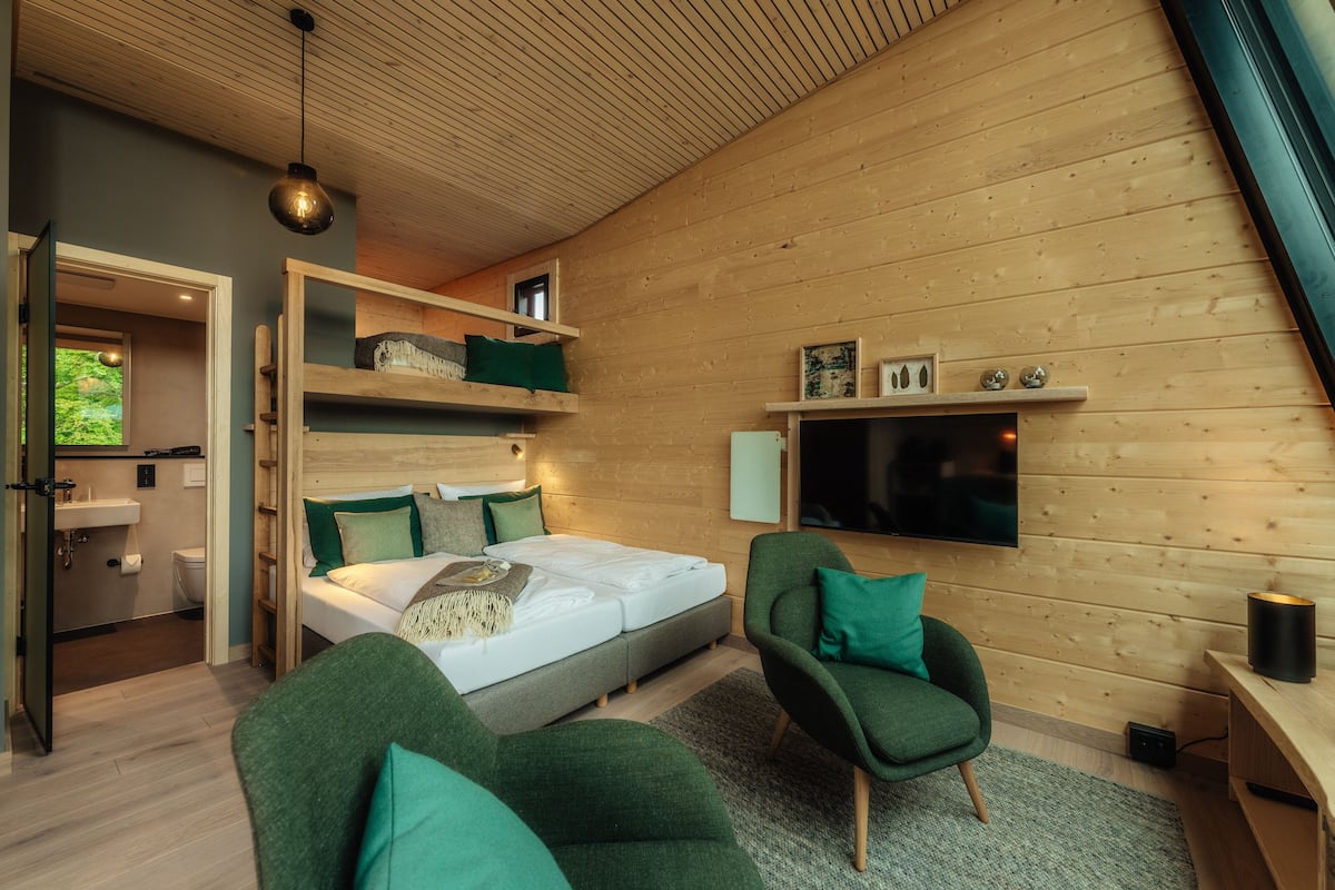 Un dormitorio moderno con una cama, dos sillas verdes, un televisor y techo de madera.