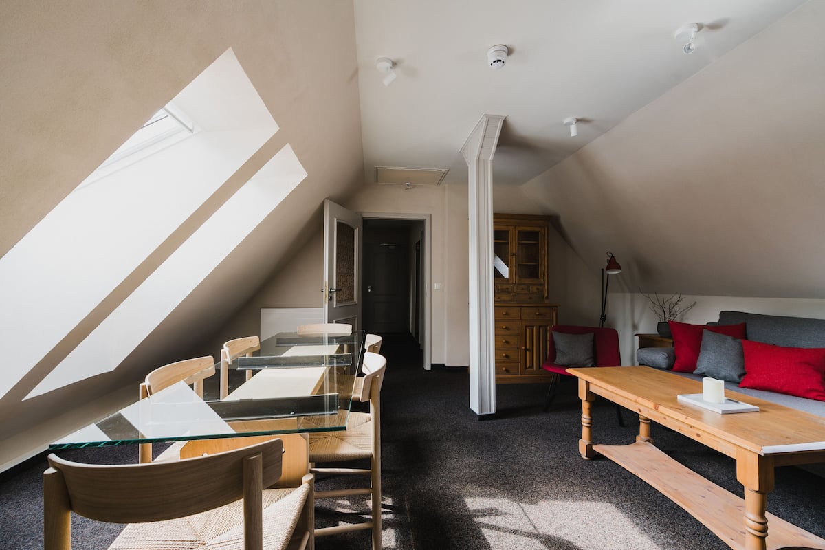 Un dormitorio tipo loft moderno con techo inclinado, una mesa de comedor de vidrio, muebles de madera y una zona de estar acogedora con cojines rojos.
