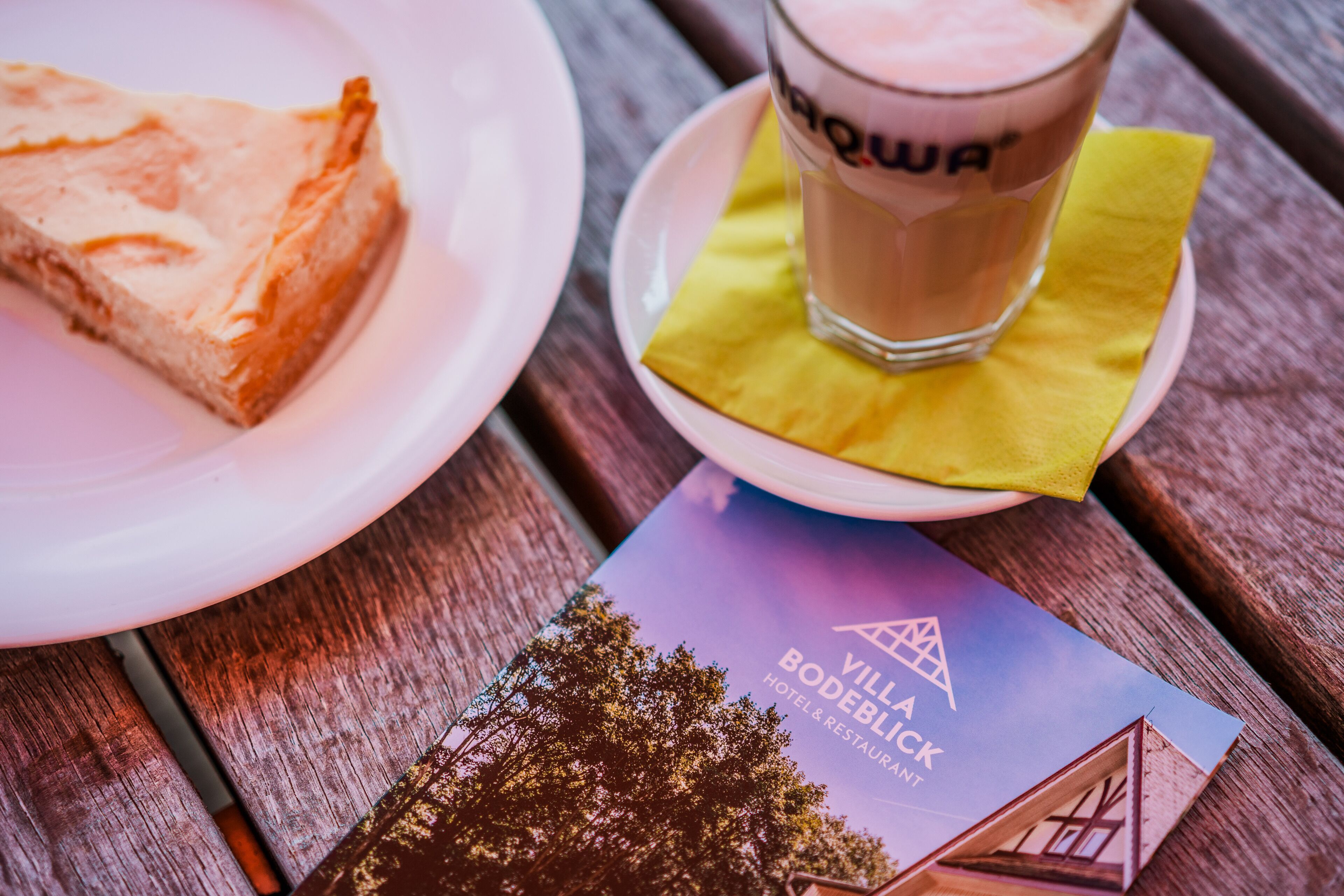 A slice of cheesecake on a white plate, a glass of beverage with a straw, and a brochure for Villa Bodeblick Hotel & Restaurant on a wooden table.