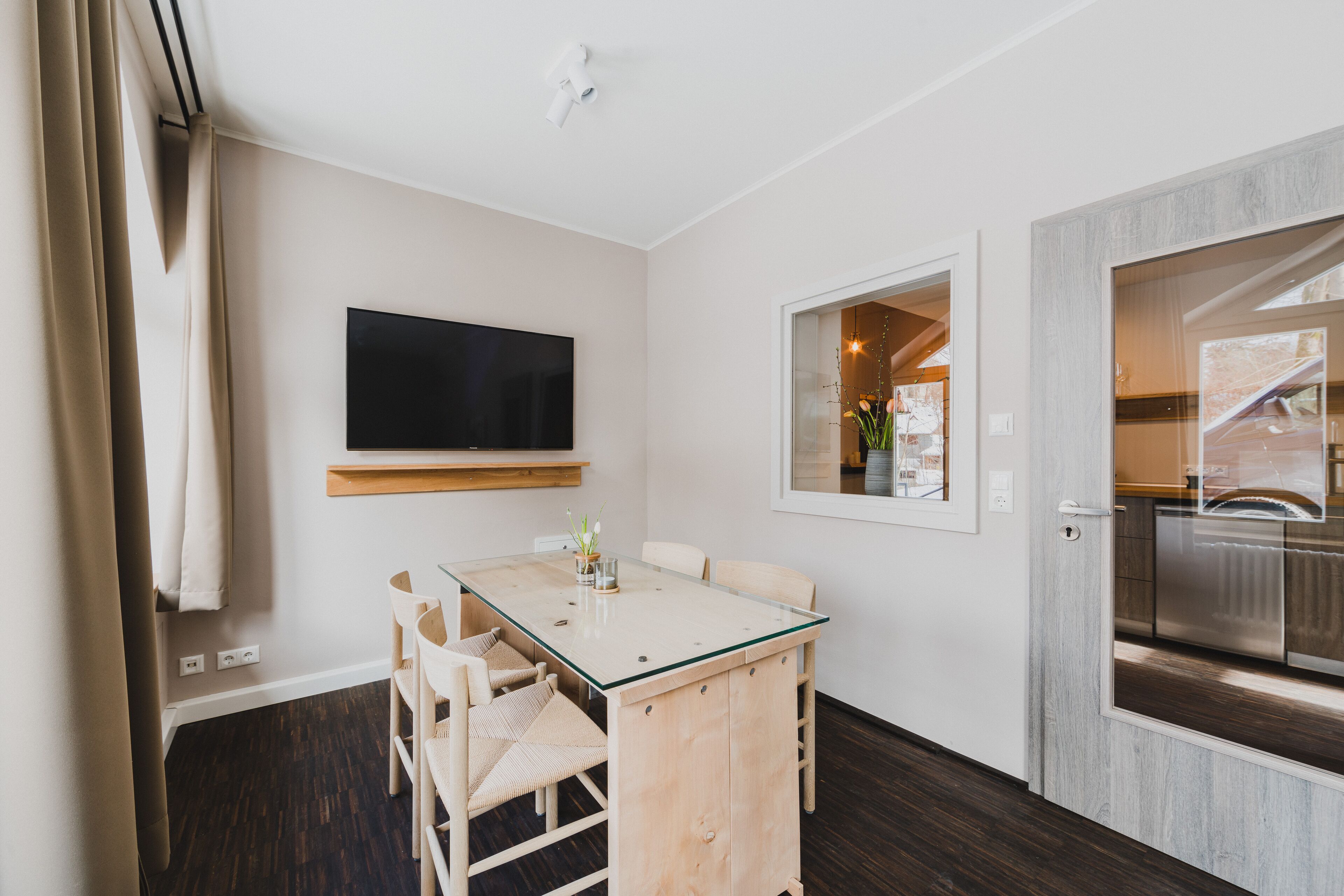 A modern kitchen with a built-in oven, a dining area with a glass-top table and chairs, and a large flat-screen TV mounted on the wall.