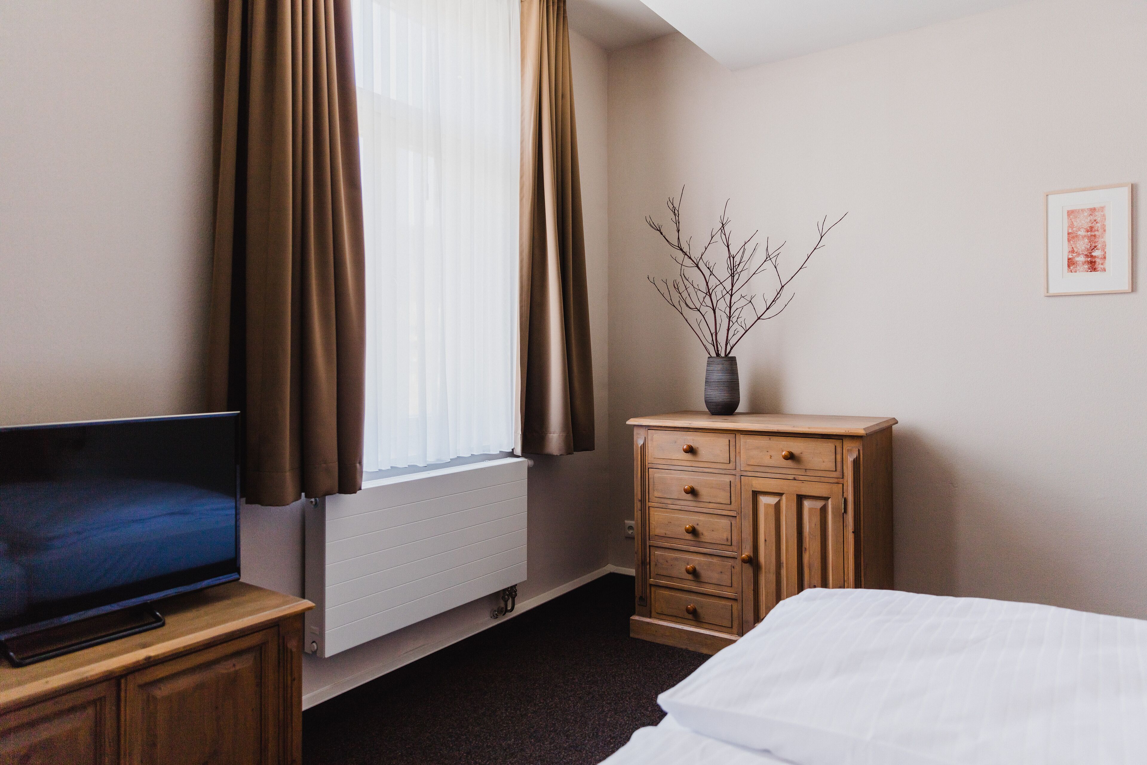 A hotel room with a bed, a TV on a wooden cabinet, a dresser, a vase with branches, and a framed picture on the wall.