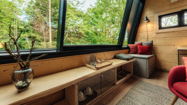 A cozy cabin interior with a large window overlooking a forest, a wooden bench, and a red chair. - Hotel Villa Bodeblick (Wernigerode)