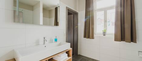 A modern bathroom with a large mirror, white sink, wooden vanity, and a window with curtains.