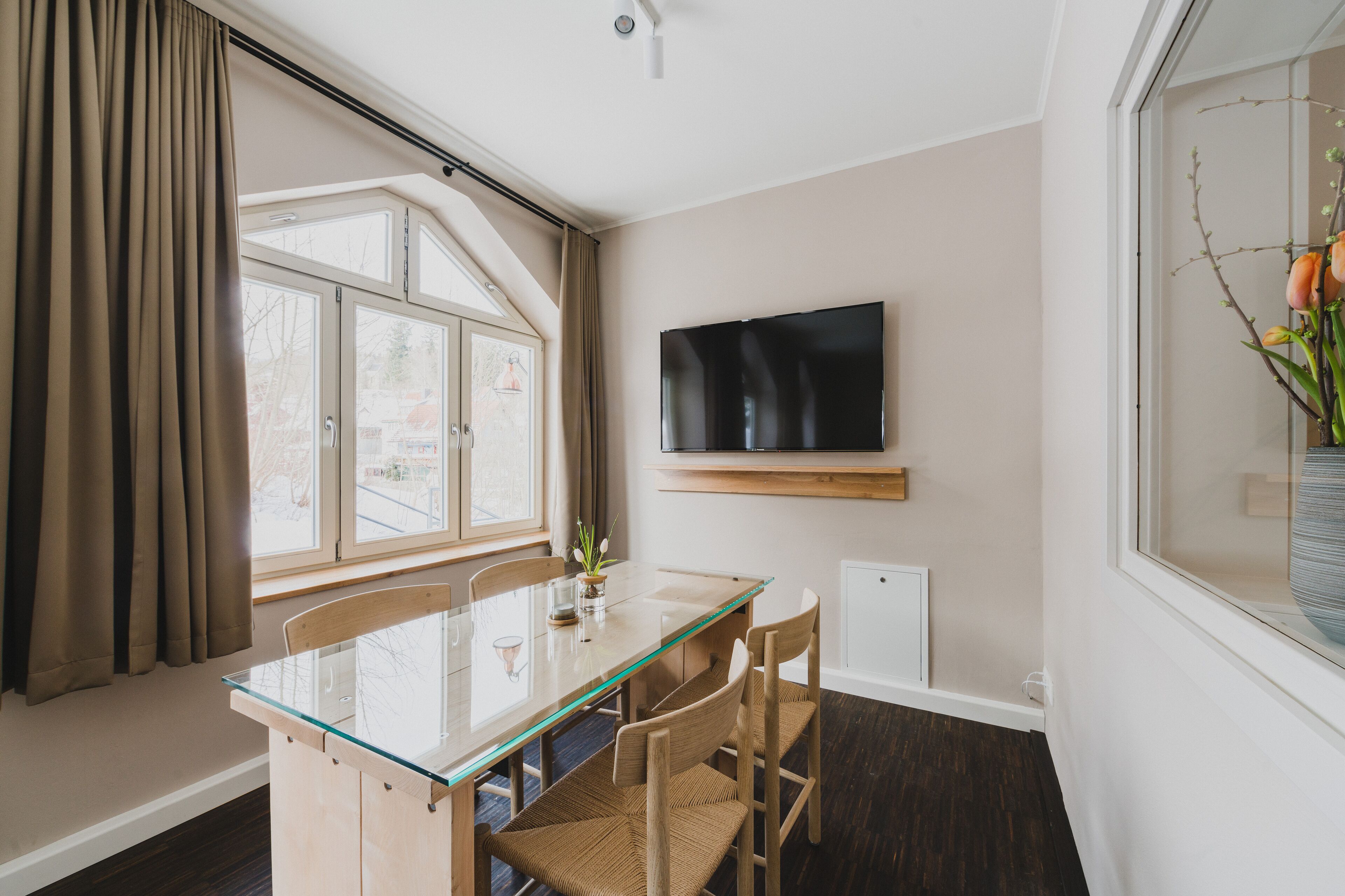 Un comedor moderno con una mesa de vidrio, sillas de madera, un televisor de pantalla plana en la pared y una ventana con cortinas.