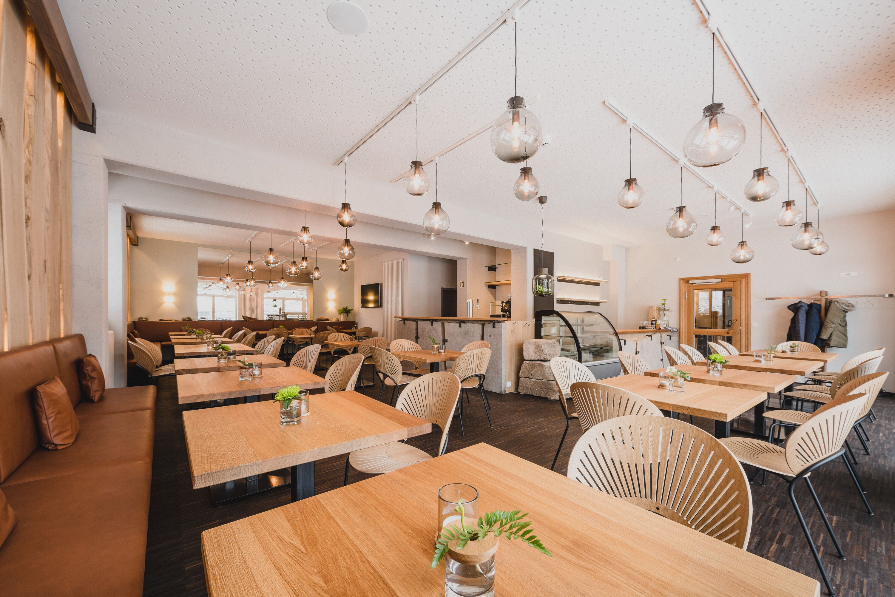 Een modern restaurant met houten tafels en stoelen, hangende吊灯 en een gezellig zitgedeelte met kussens.