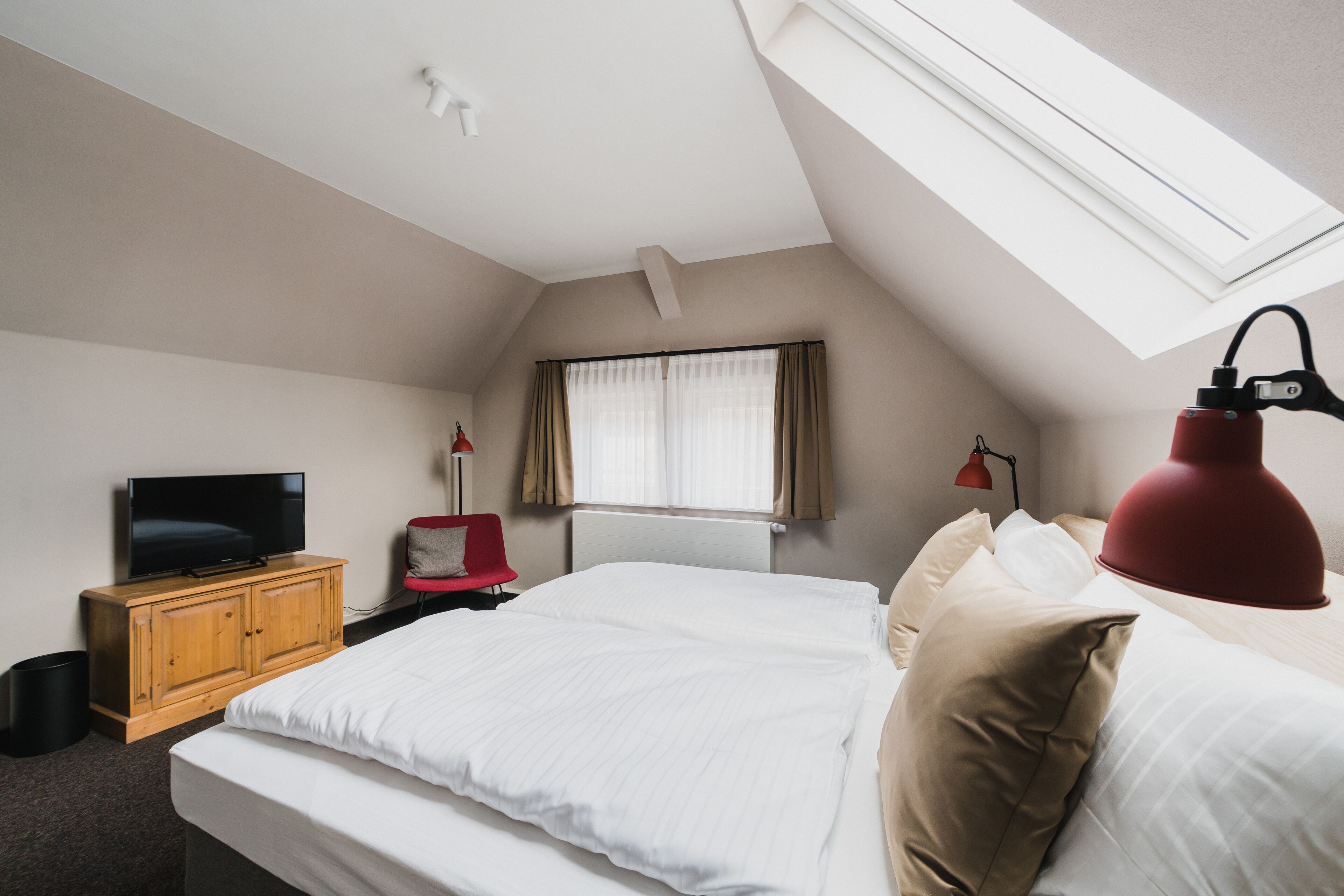 Un dormitorio con cama, televisión sobre un mueble de madera, ventana con cortinas y tragaluz.