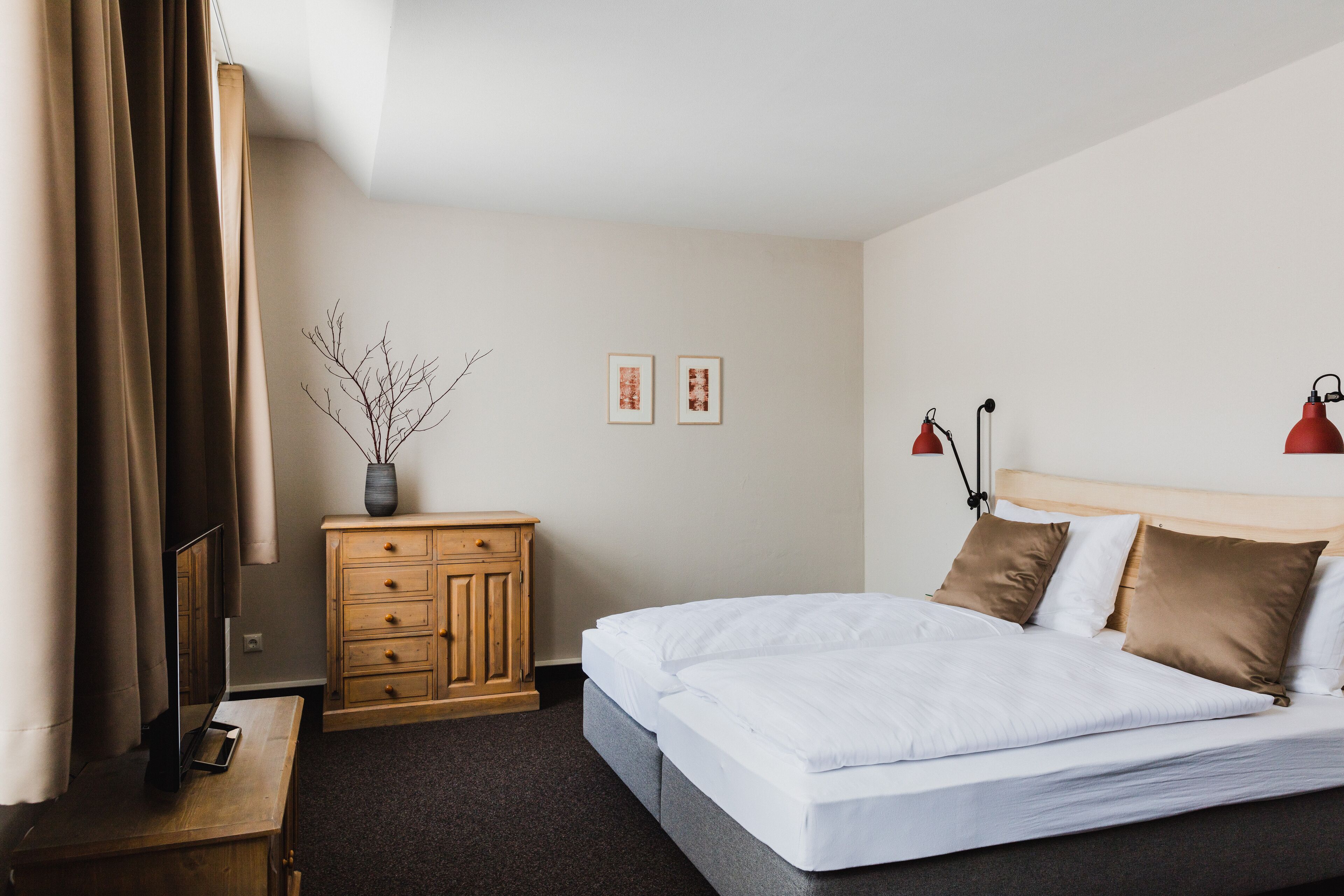 A hotel room with a bed, wooden dresser, two framed pictures, a vase with branches, and two red lamps.