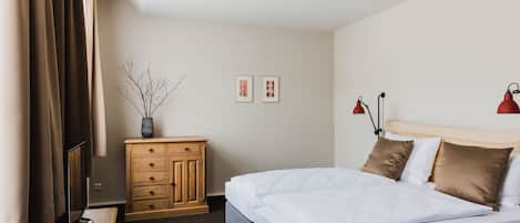 A hotel room with a bed, wooden dresser, two framed pictures, a vase with branches, and two red lamps.