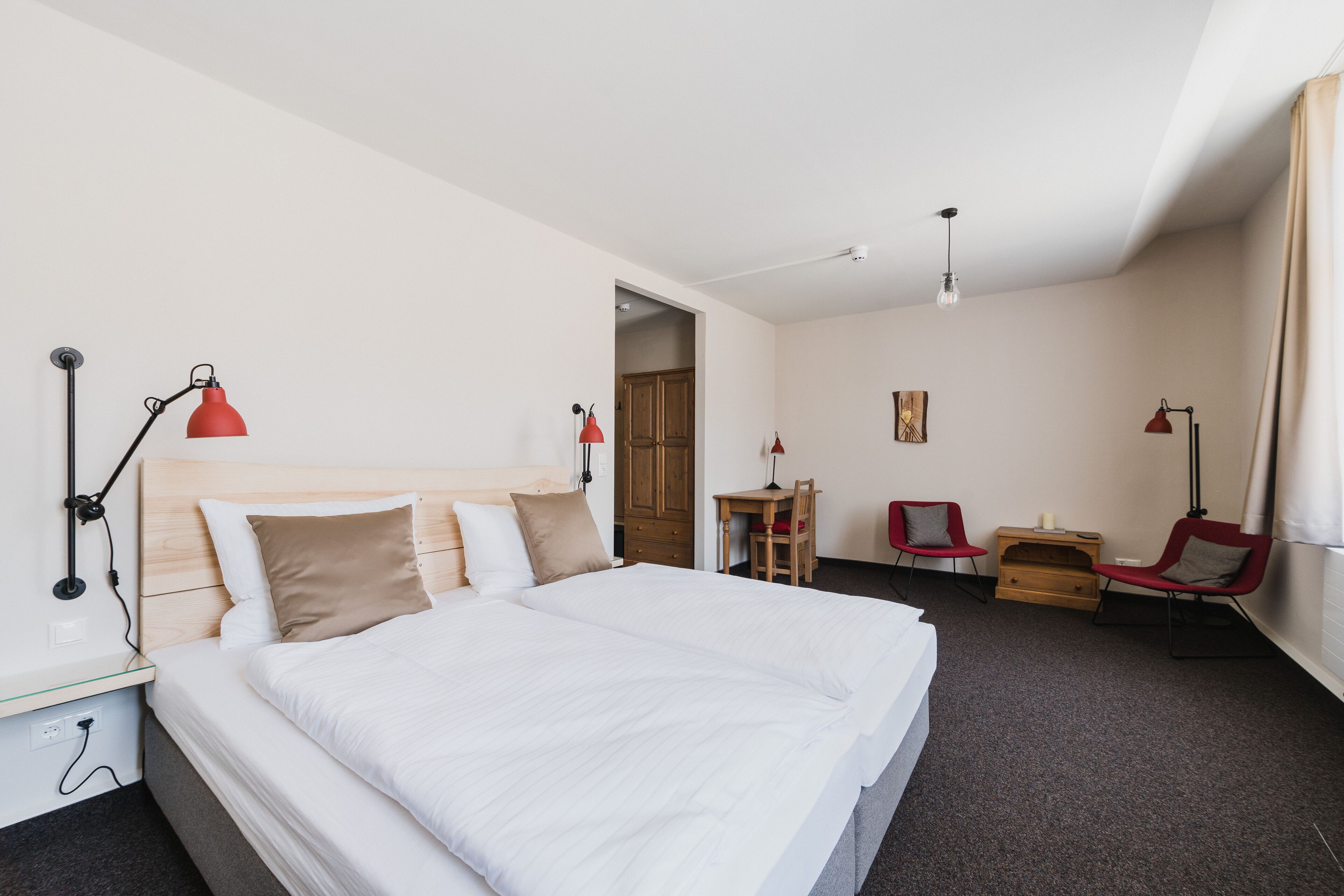 Une chambre d’hôtel avec un grand lit, deux fauteuils rouges, un bureau et une armoire en bois.