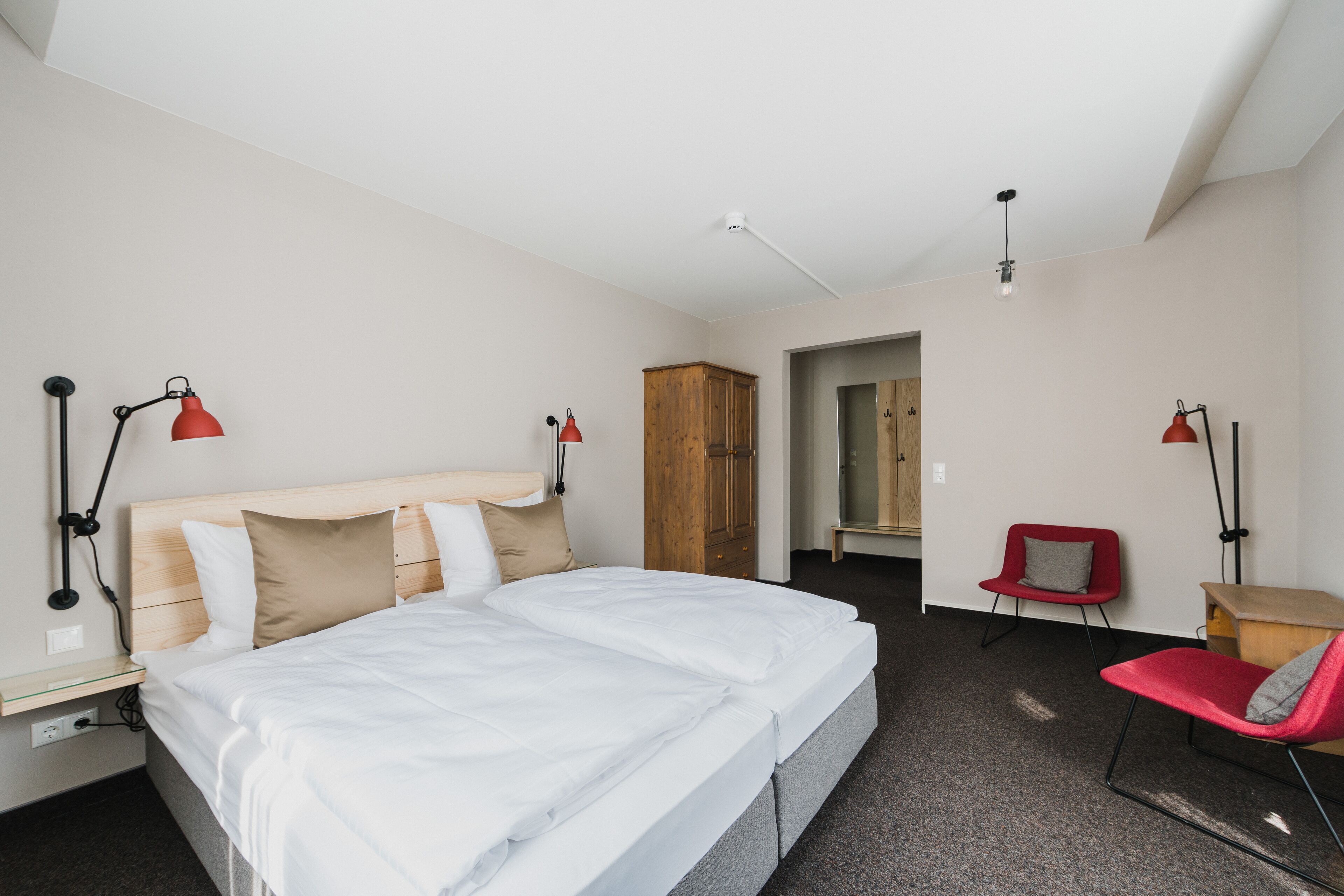 A modern hotel room with a large bed, two red chairs, a wooden wardrobe, and a small desk.